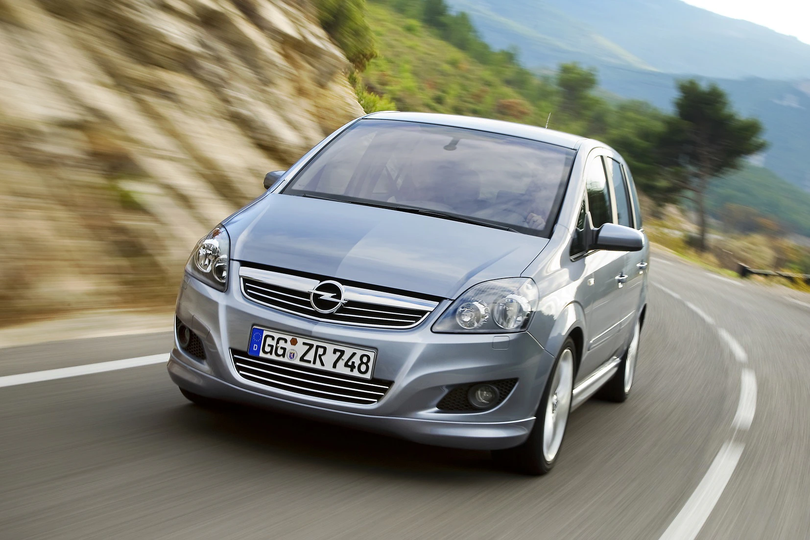 Silberner Opel Van fährt auf kurviger Bergstraße, umgeben von grüner Landschaft und Felsen im Hintergrund.