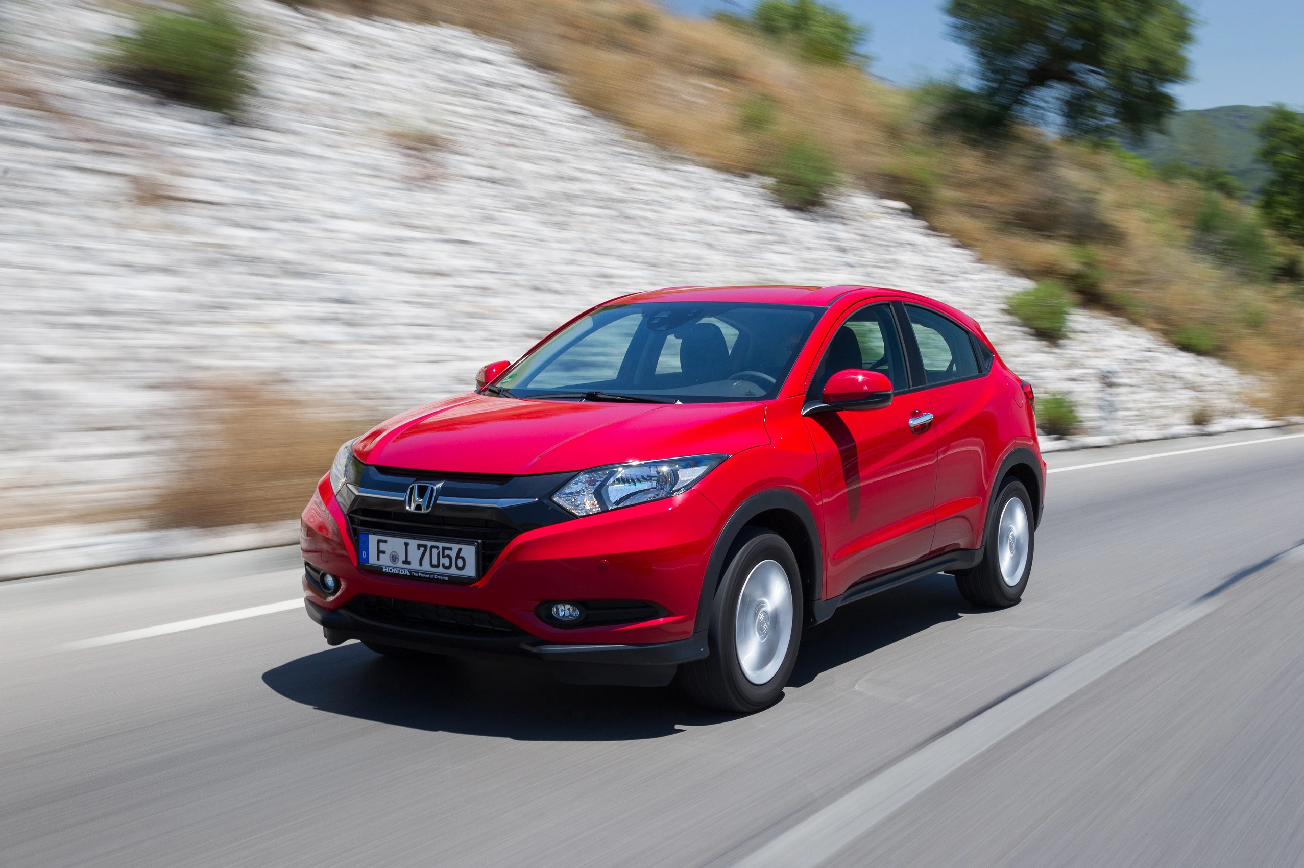 Roter SUV fährt auf einer Landstraße entlang einer Steinmauer, bei sonnigem Wetter und grüner Umgebung im Hintergrund.