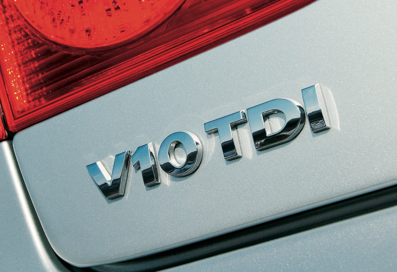Nahaufnahme eines silbernen Autos mit dem Emblem "V10 TDI" in Chromoptik auf der Heckklappe. Rotes Rücklicht im Hintergrund.