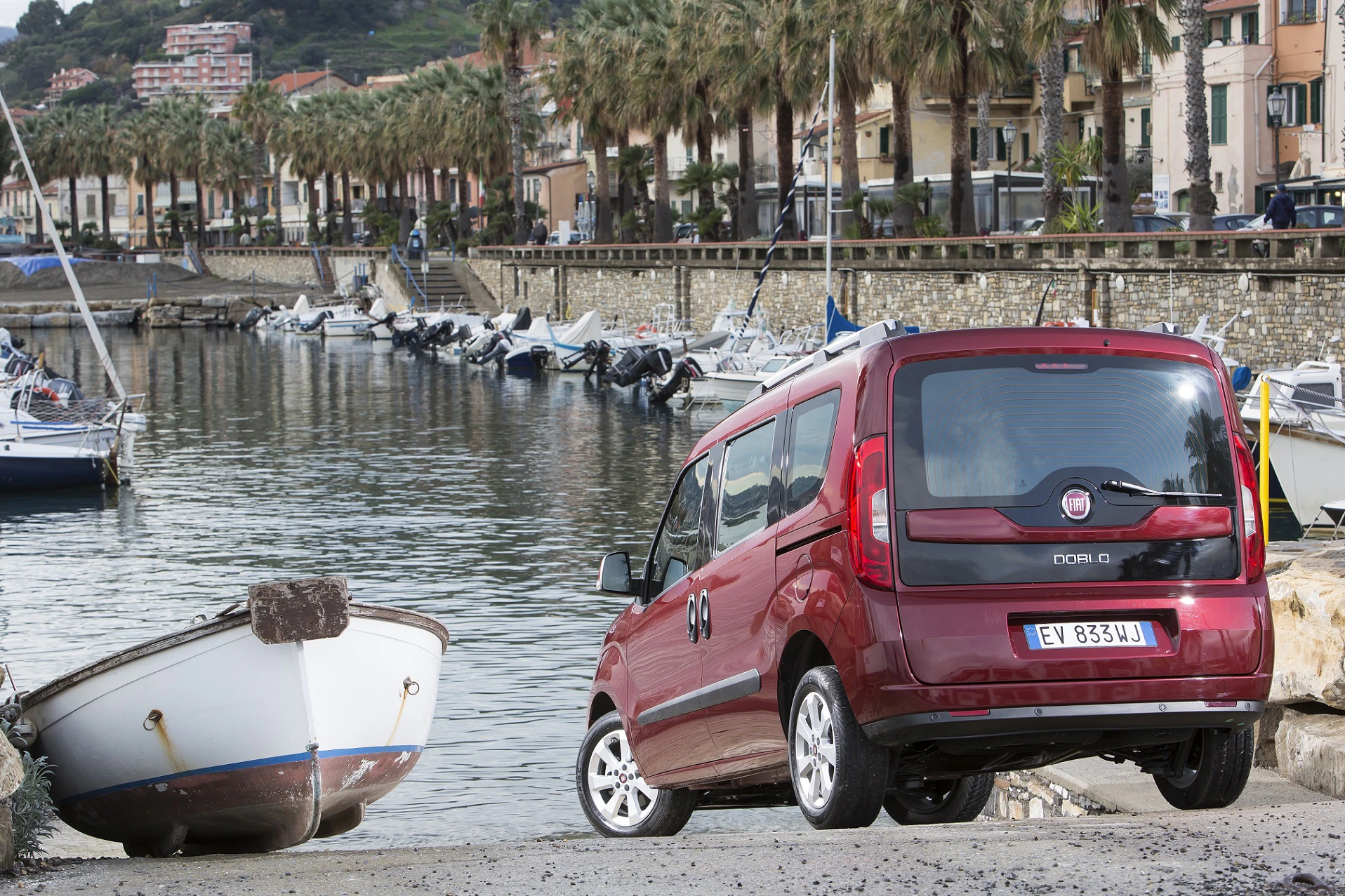 Ein roter Minivan parkt am Wasser neben einem Boot. Im Hintergrund sind Palmen und eine Reihe von Booten vor einer Stadtlandschaft.
