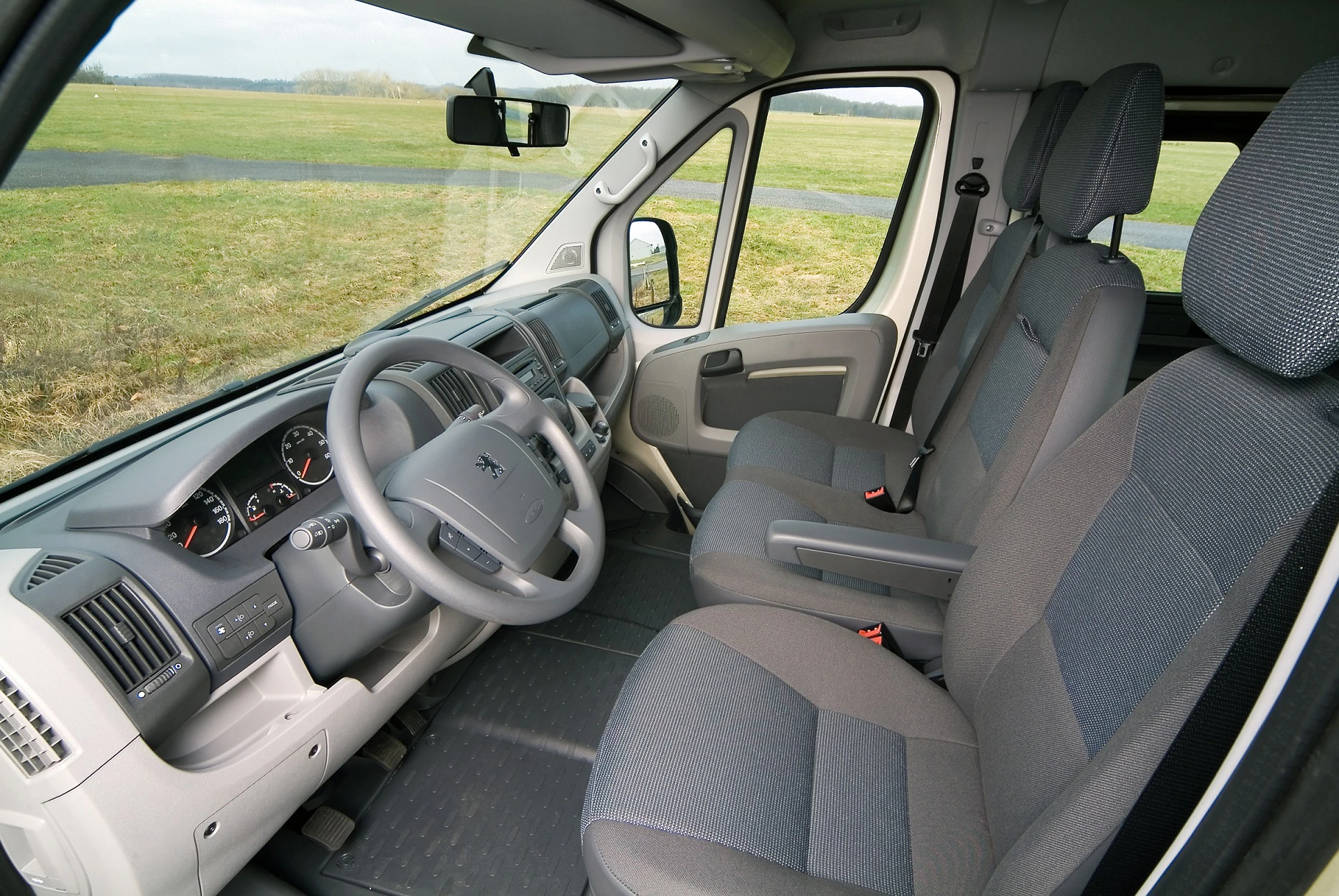 Innenraum eines Transporters mit grauem Interieur, zwei Vordersitzen, Lenkrad und Armaturenbrett. Draußen ist eine grüne Wiese sichtbar.