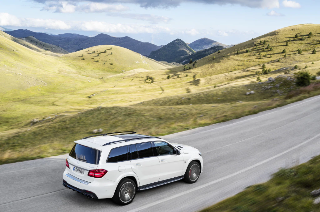 Weißer SUV fährt auf einer asphaltierten Straße durch hügelige Landschaft mit grünen Wiesen und Bergen im Hintergrund unter blauem Himmel.