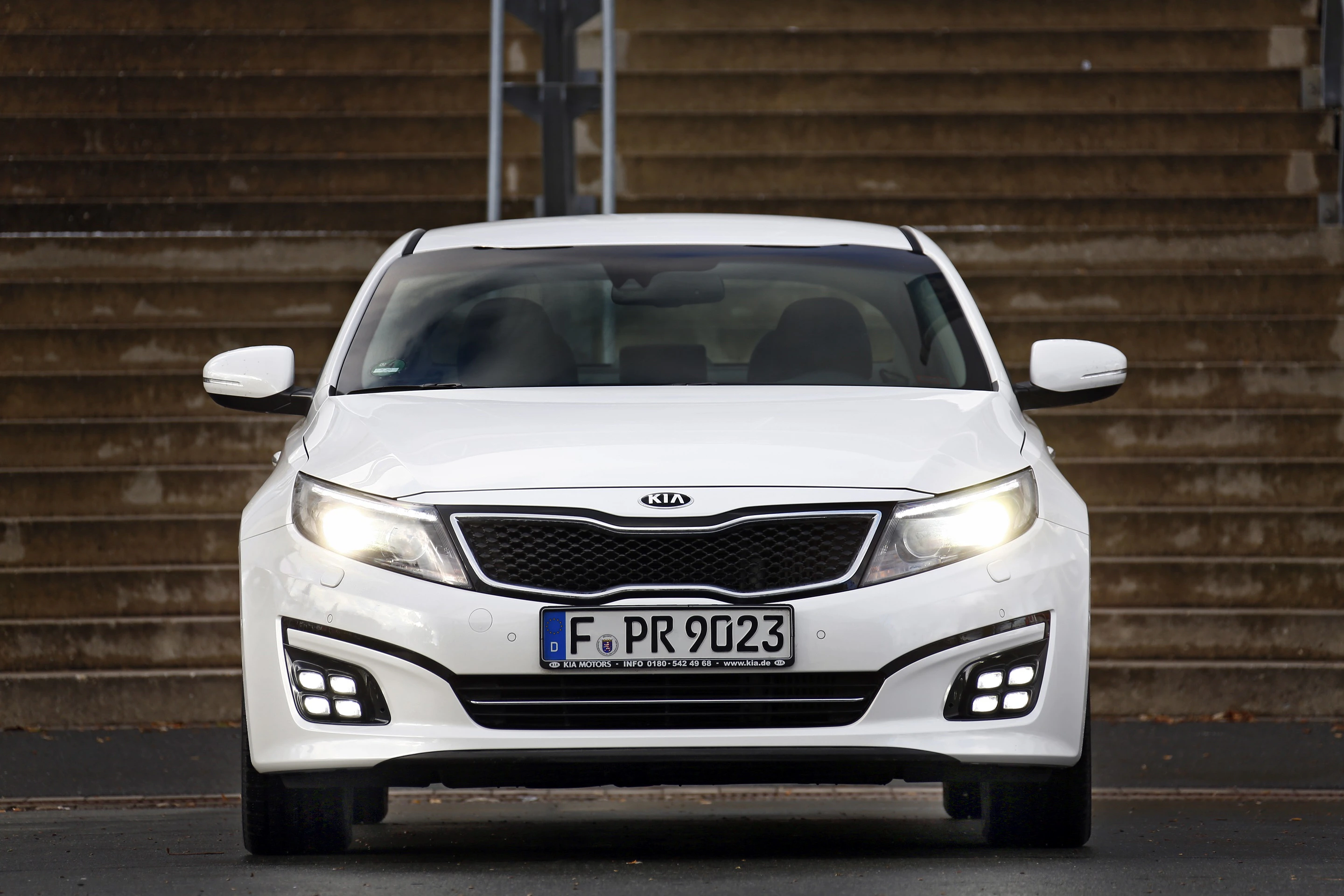 Weißer Kia Optima steht frontal vor Treppe; eingeschaltete LED-Tagfahrlichter; deutsches Nummernschild.