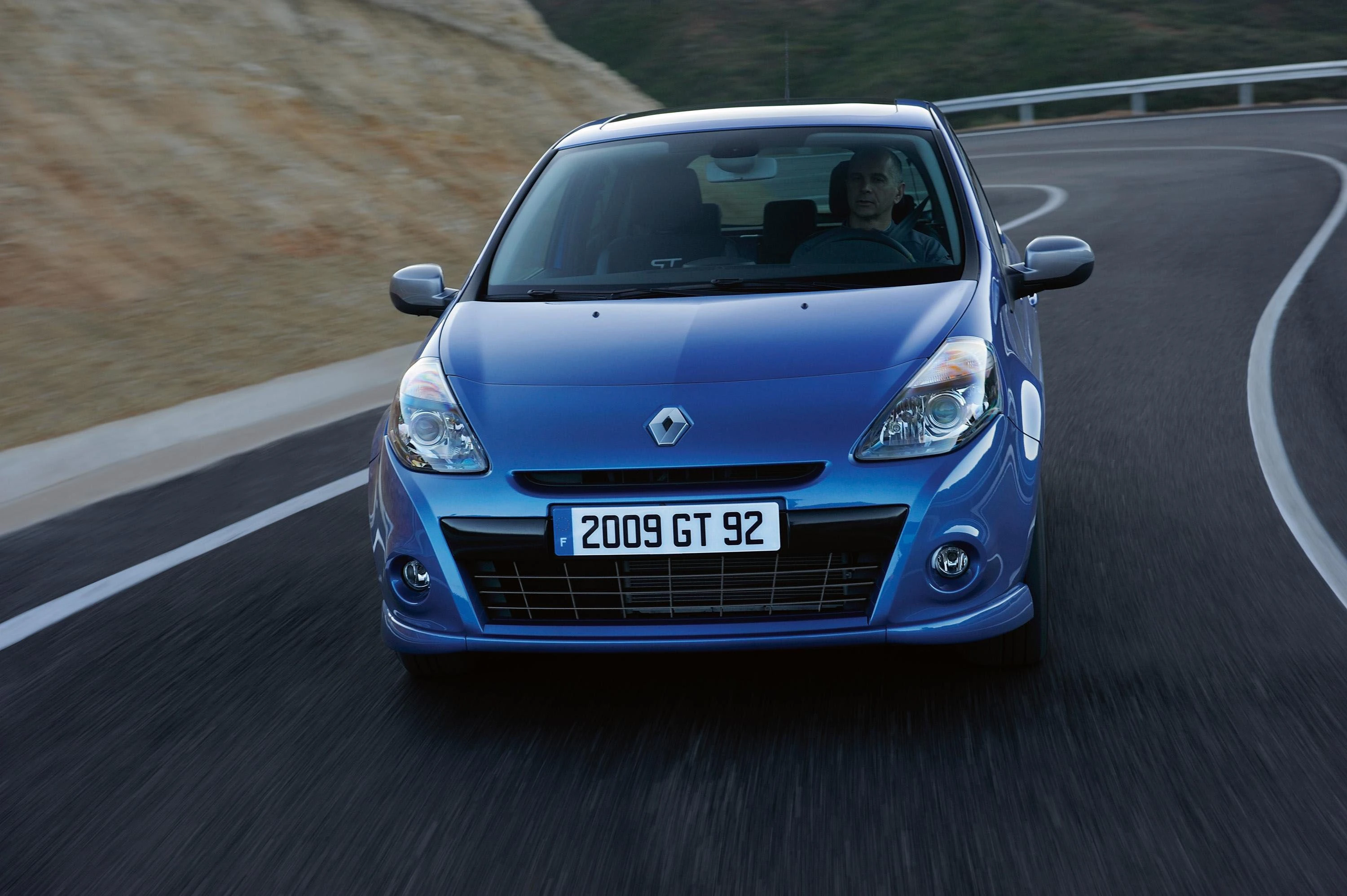 Blaues Auto, frontal fotografiert, fährt auf einer kurvigen Straße. Fahrer sichtbar, Nummernschild zeigt "2009 GT 92".