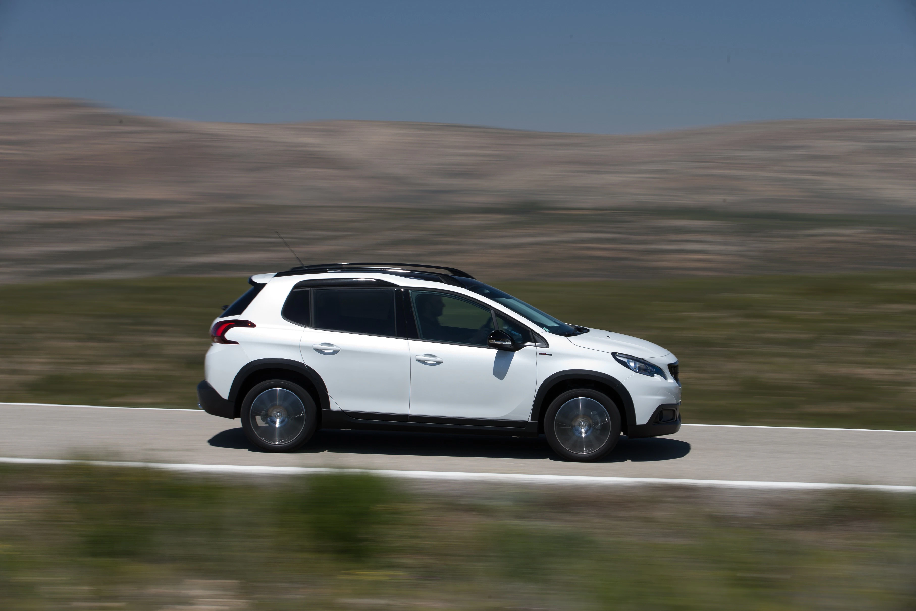 Weißer SUV fährt schnell auf einer Landstraße; unscharfer Hintergrund mit Hügeln und blauem Himmel.