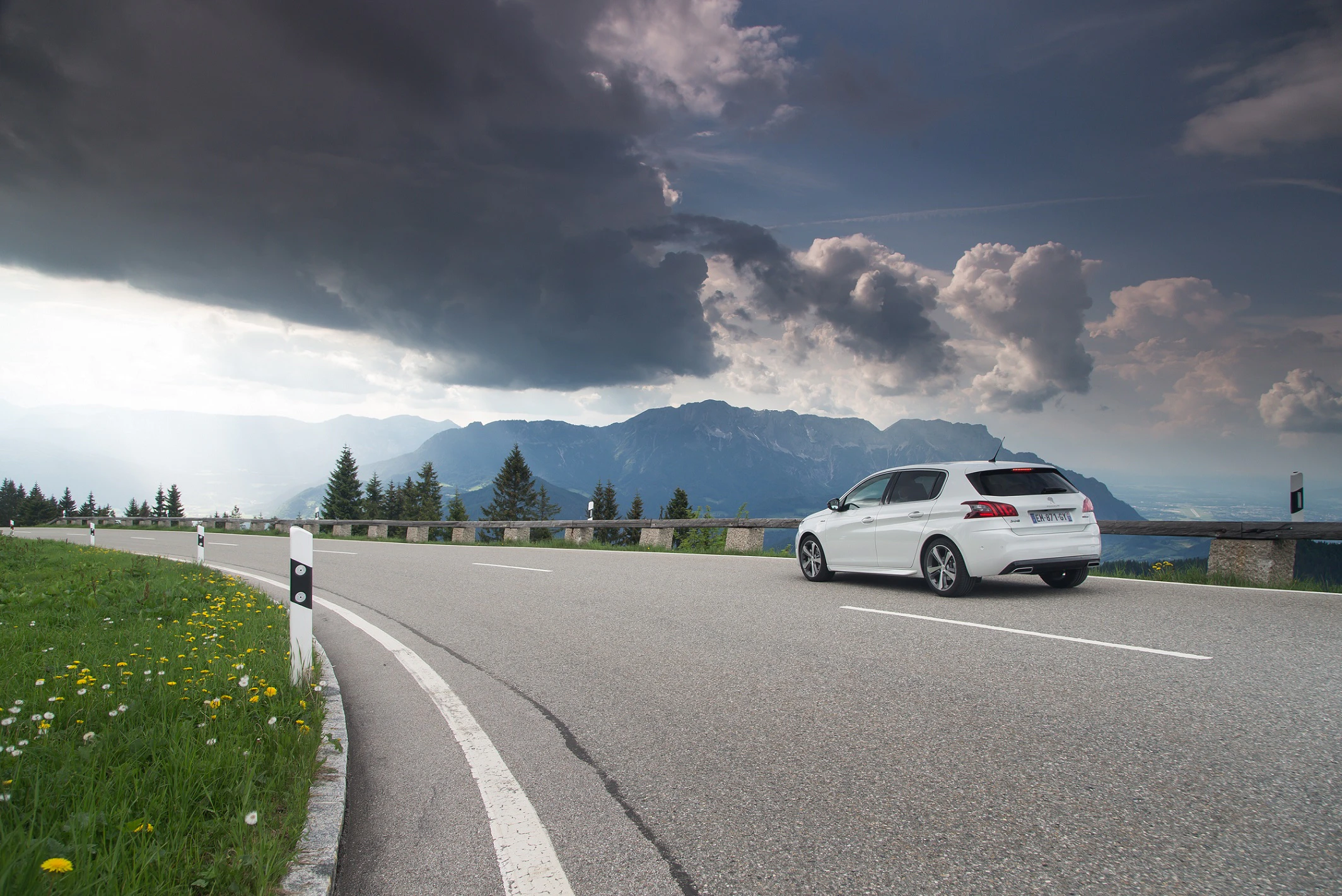 Weißes Auto fährt auf kurviger Bergstraße, umgeben von Wiesen und Bergen. Dramatische Wolkenformationen am Himmel.