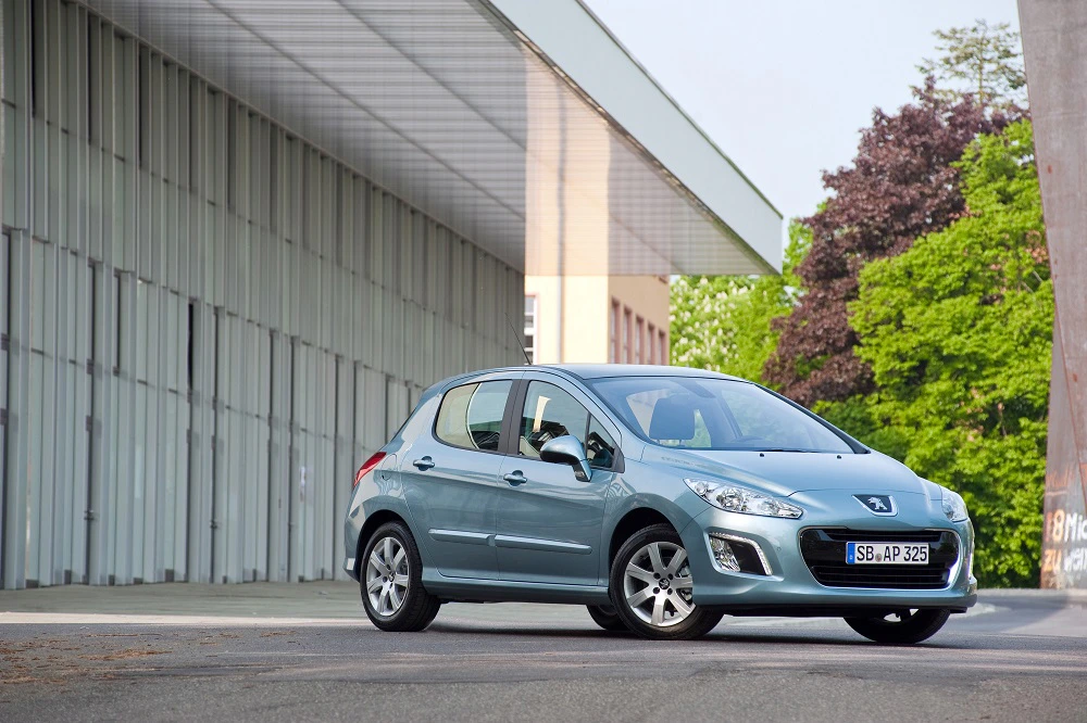 Silberner Peugeot 308 steht vor einer großen, modernen Lagerhalle mit grünen Bäumen im Hintergrund.