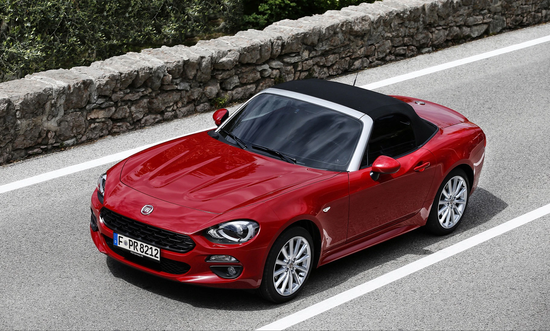 Roter Cabrio auf einer Straße neben einer Steinmauer, mit schwarzem Dach und glänzender Lackierung.