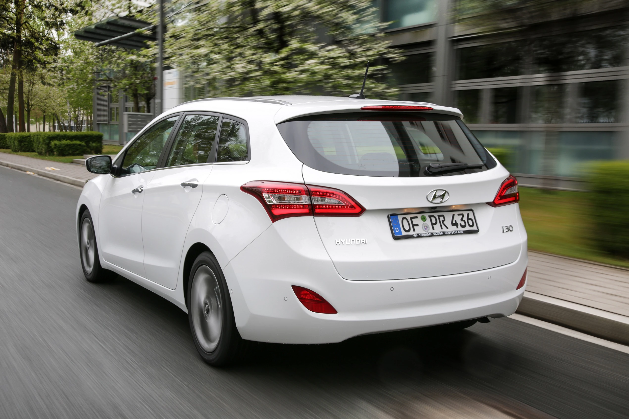 Weißer Hyundai i30 fährt auf einer Straße, umgeben von Grünen und modernen Gebäuden. Rückansicht des Fahrzeugs bei Tageslicht.