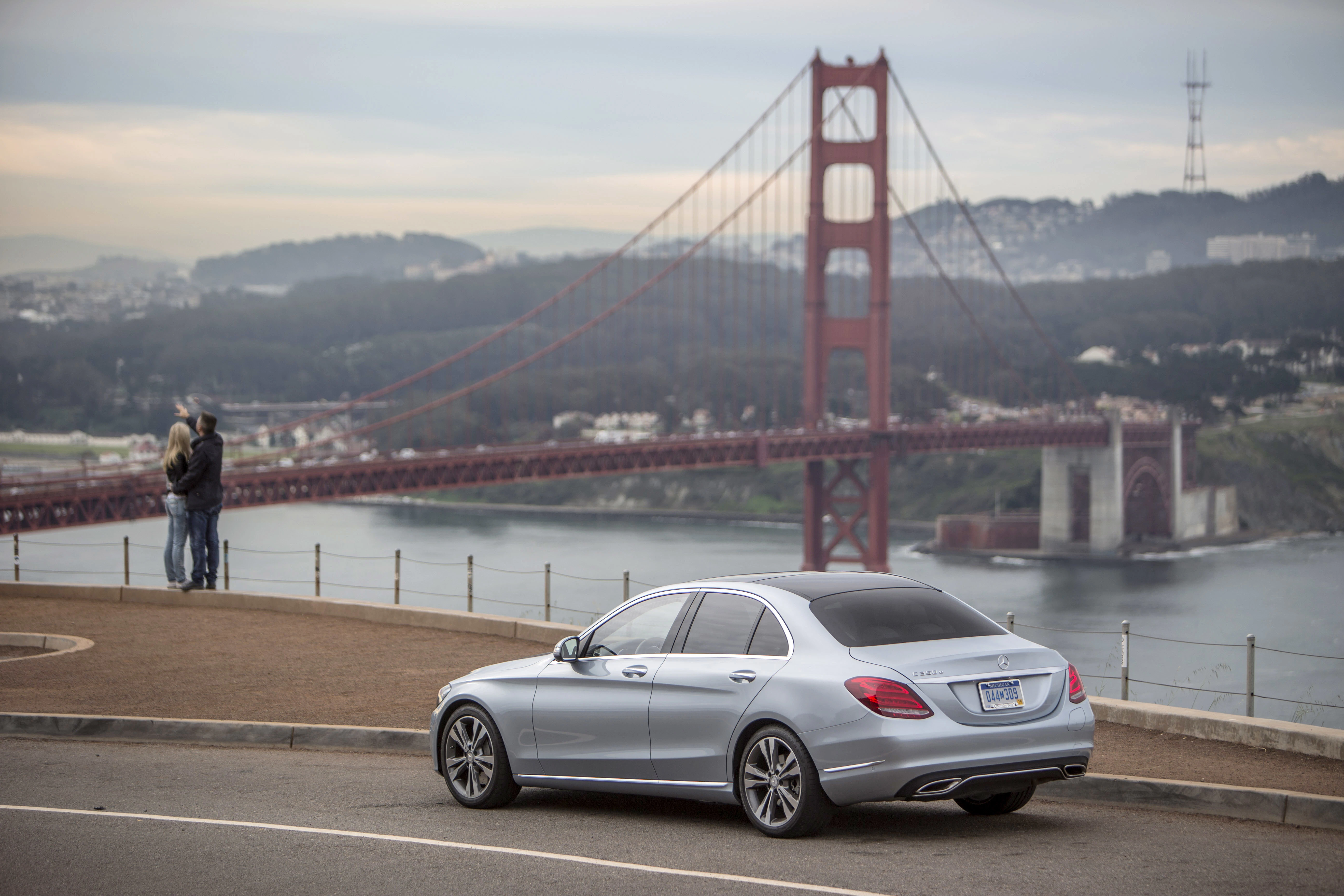 Silbernes Auto auf einer Anhöhe, im Hintergrund die Golden Gate Bridge und verschwommene Skyline. Ein Paar steht an der Absperrung und betrachtet die Aussicht.