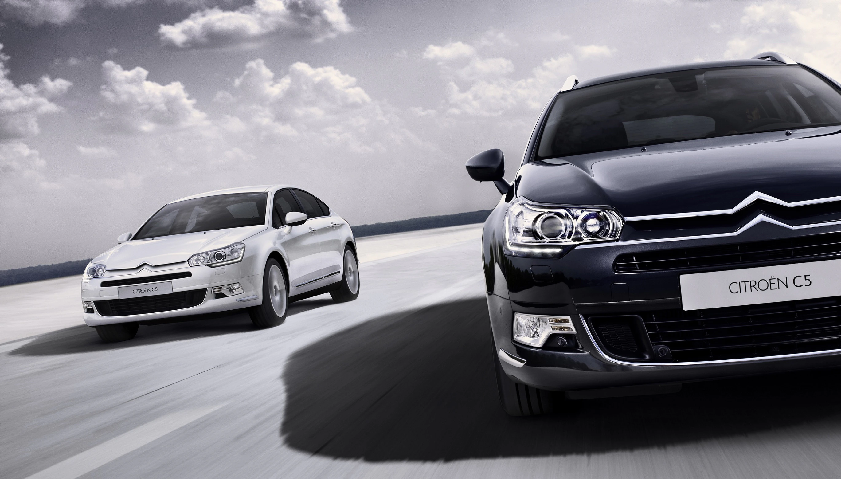Zwei Citroën C5-Fahrzeuge fahren auf einer offenen Straße unter bewölktem Himmel, ein schwarzes im Vordergrund und ein weißes im Hintergrund.