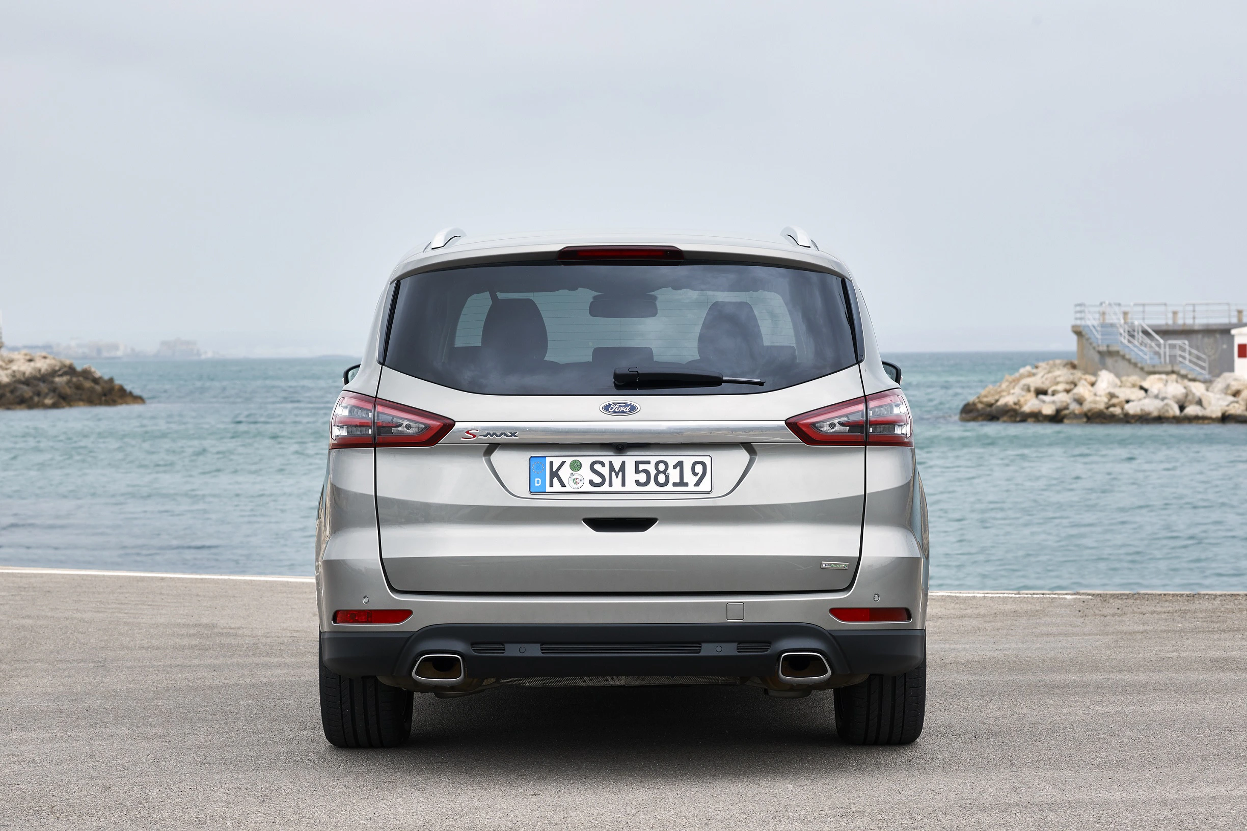 Silberner Ford S-MAX, Heckansicht, parkt auf einer Straße am Meer. Himmel bewölkt, Wasser und Felsen im Hintergrund.