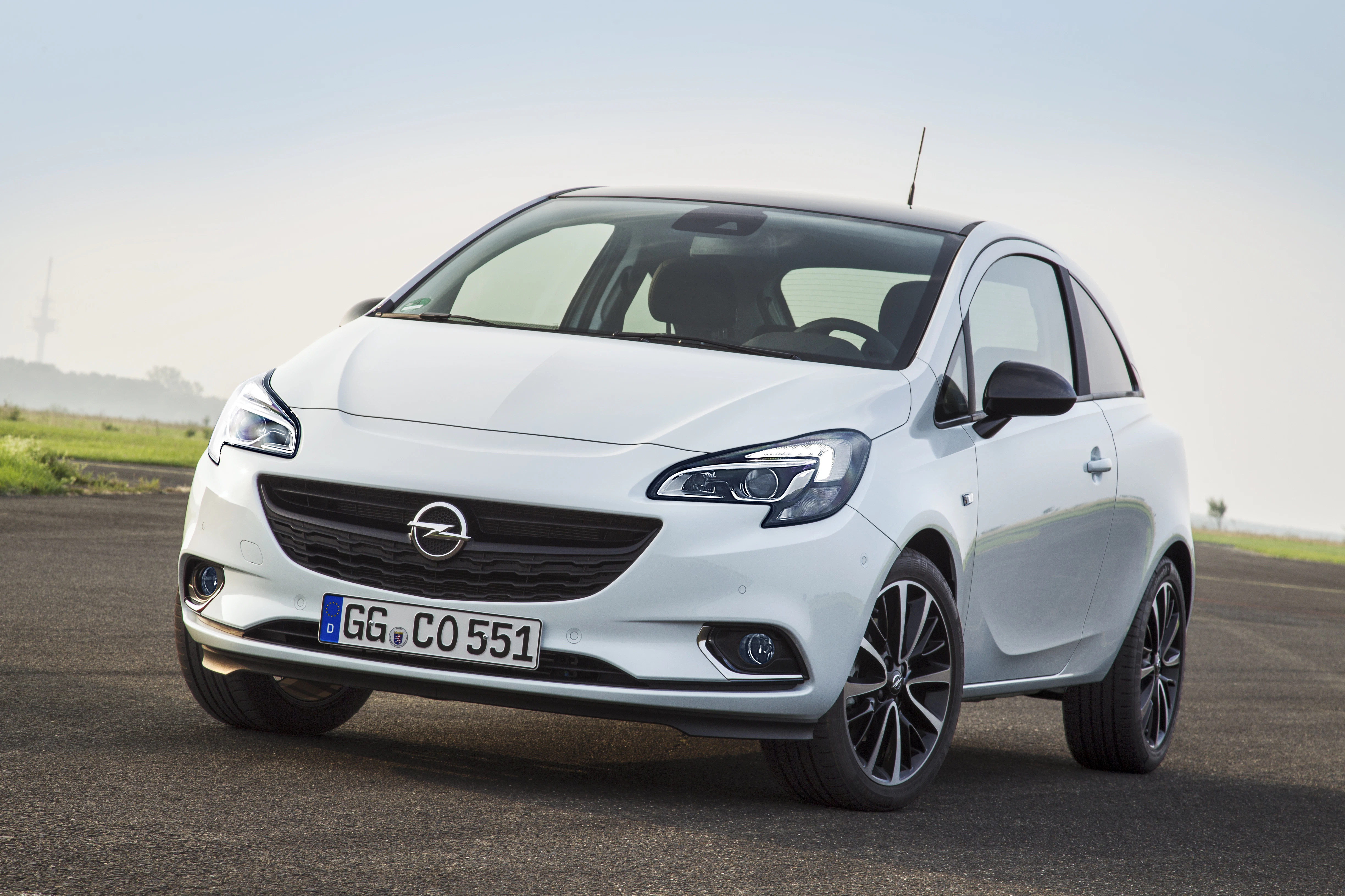 Weißer Opel Corsa auf einer Straße, leicht schräg von vorne fotografiert, mit schwarzem Dach und modernem Design vor wolkigem Himmel.