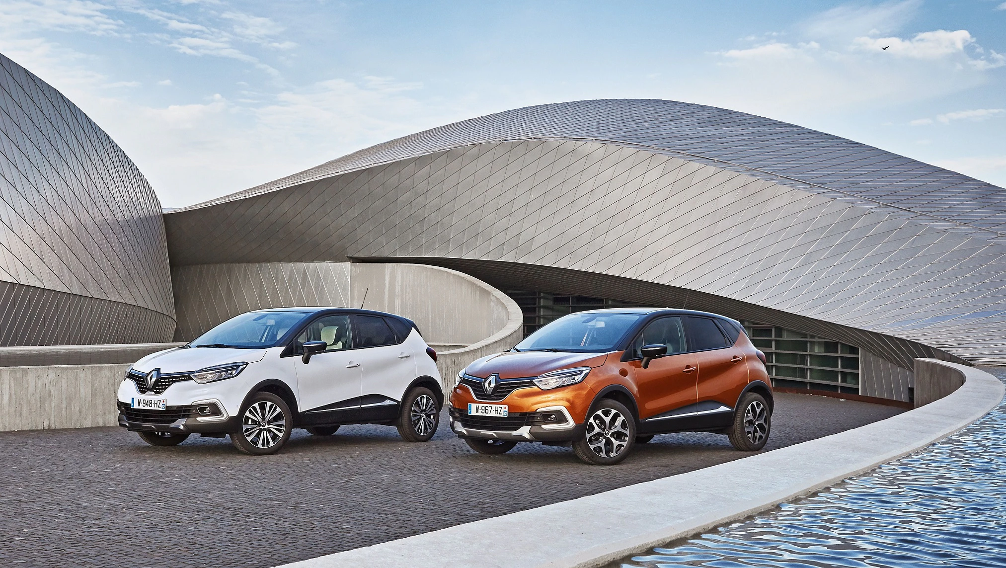 Zwei Renault-SUVs, einer weiß und einer orange, stehen vor einem modernen, geschwungenen Gebäude bei sonnigem Wetter.