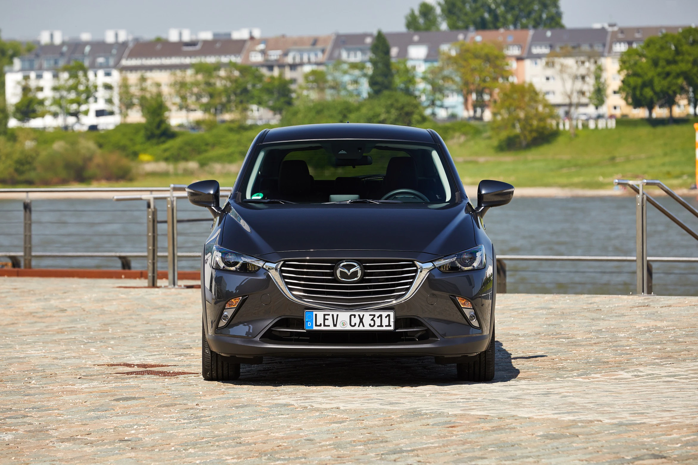 Schwarzer Mazda CX-3 steht auf einem gepflasterten Platz vor einem Fluss, im Hintergrund Wohngebäude und Bäume.
