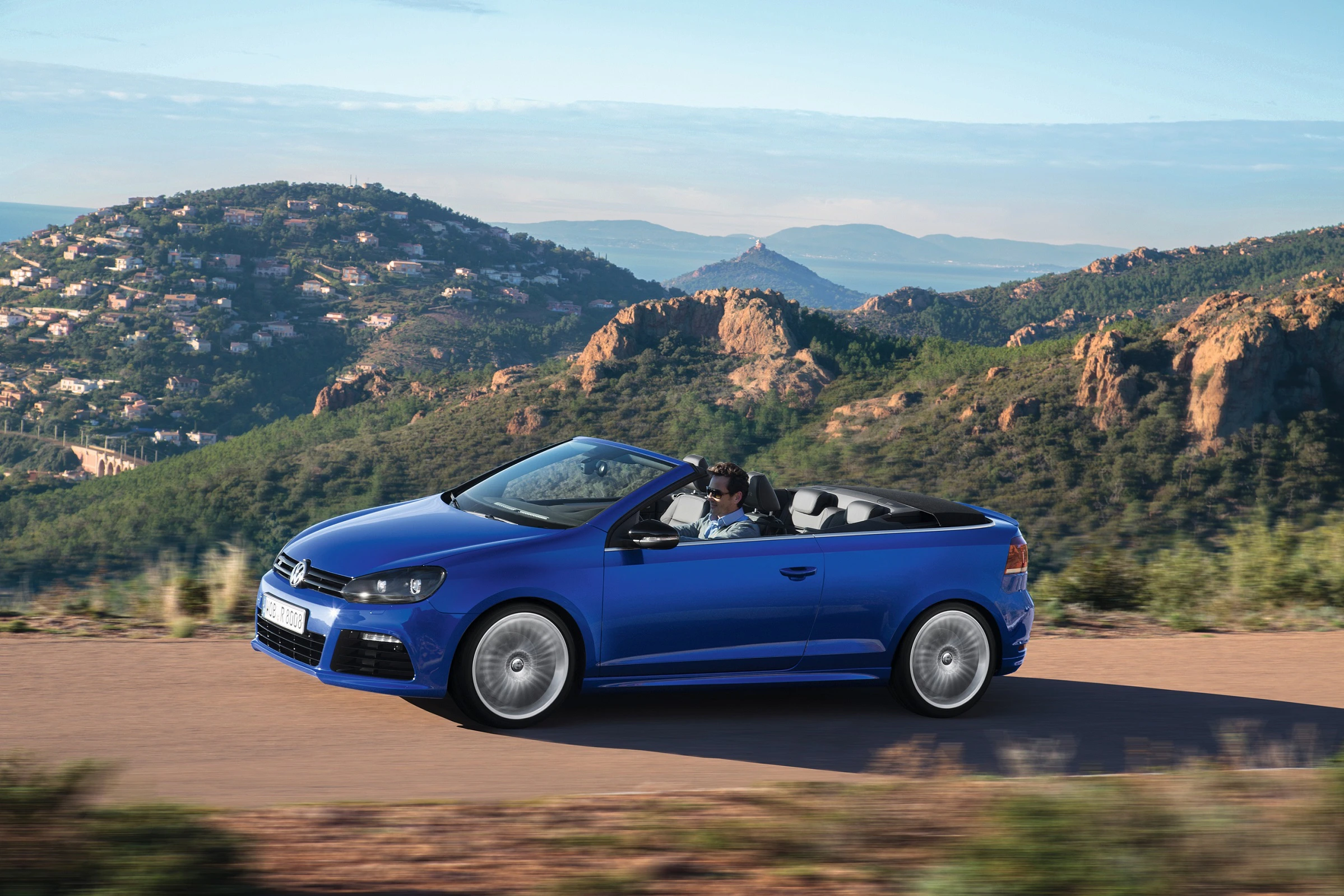 Blaues Cabriolet fährt auf einer Landstraße mit bergiger Landschaft im Hintergrund. Sonniges Wetter, klarer Himmel.