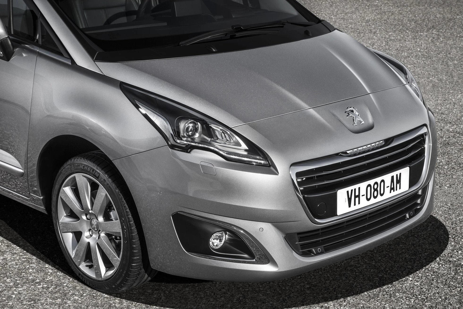 Silberner Peugeot mit glänzendem Finish, Frontansicht, markante Scheinwerfer und sichtbares Nummernschild auf grauem Untergrund.
