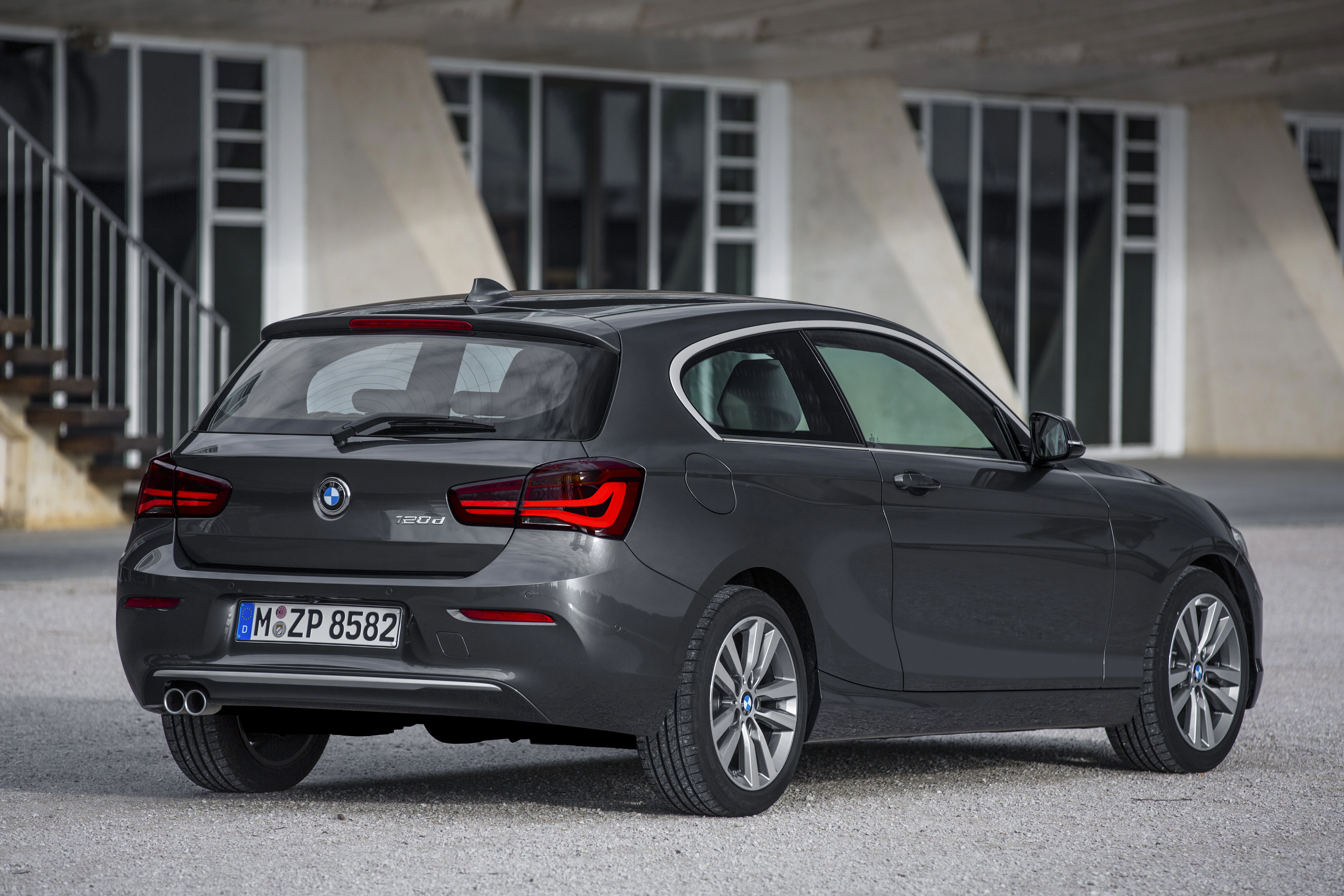 Rückansicht eines grauen BMW 1er, geparkt vor einem modernen Gebäude mit Stufen und Betonpfeilern.