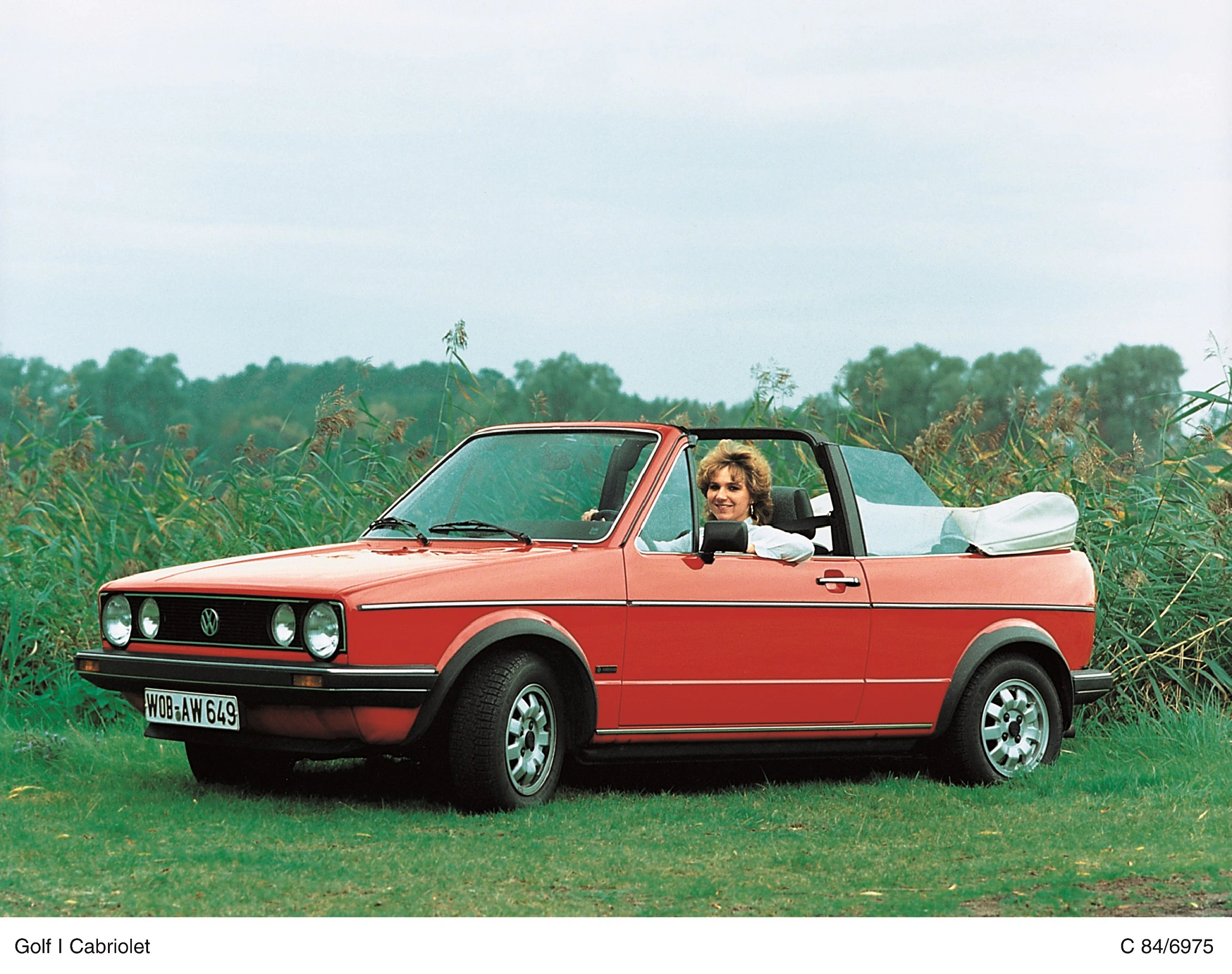 Rotes Volkswagen Golf I Cabriolet auf einer Wiese, mit offenem Verdeck. Eine Person sitzt lächelnd am Steuer.