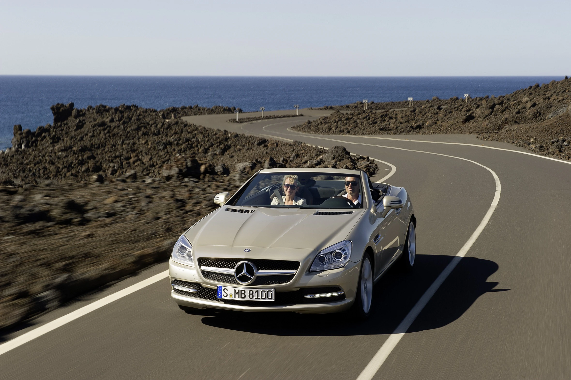 Zwei Personen fahren in einem silbernen Mercedes-Cabrio auf einer geschwungenen Küstenstraße mit Felsen und Meer im Hintergrund.