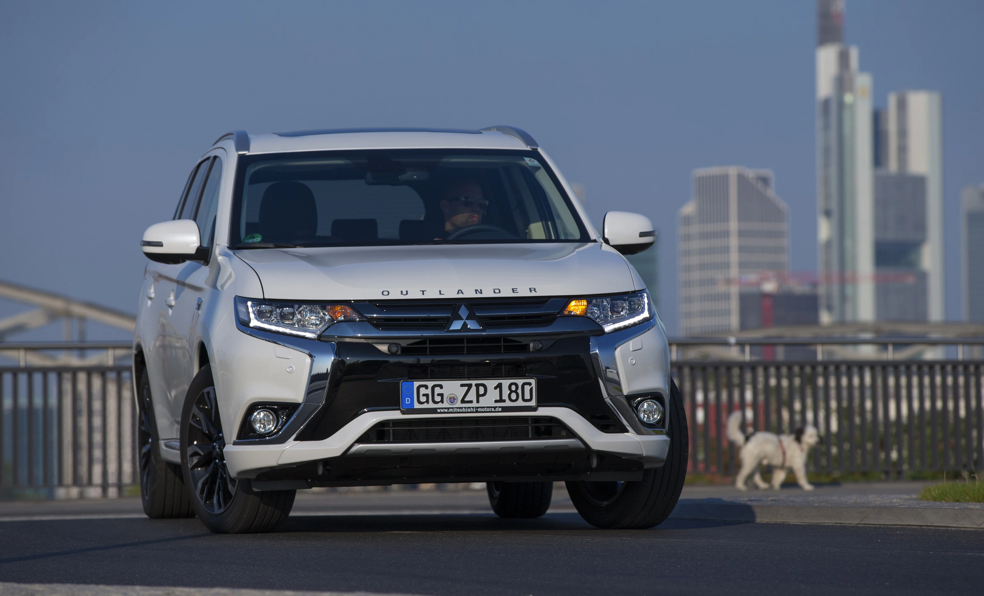 Weißer Mitsubishi Outlander fährt auf einer Straße, im Hintergrund sind Hochhäuser und ein kleiner Hund zu sehen.