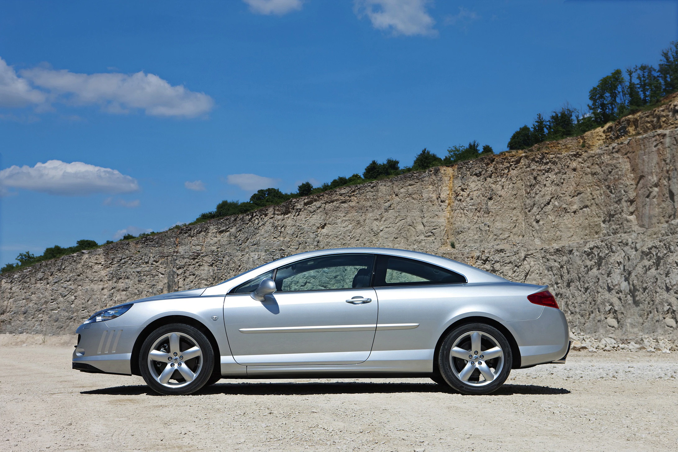 Silbernes Auto in Seitenansicht, geparkt vor einem steinigen, felsigen Hintergrund bei sonnigem Wetter mit blauem Himmel.