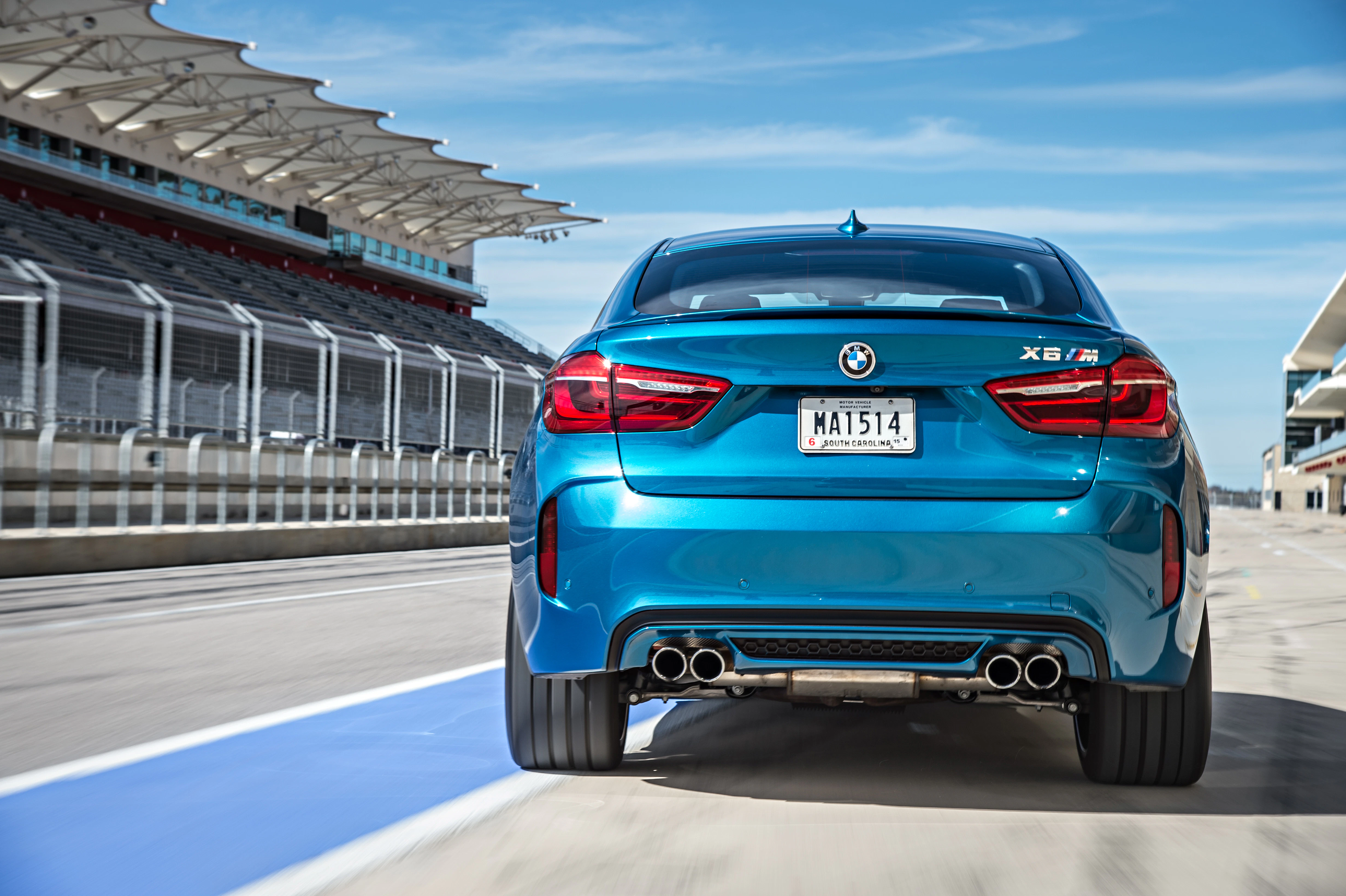 Blauer BMW X6 M von hinten auf einer Rennstrecke, umgeben von Tribünen und unter klarem Himmel.