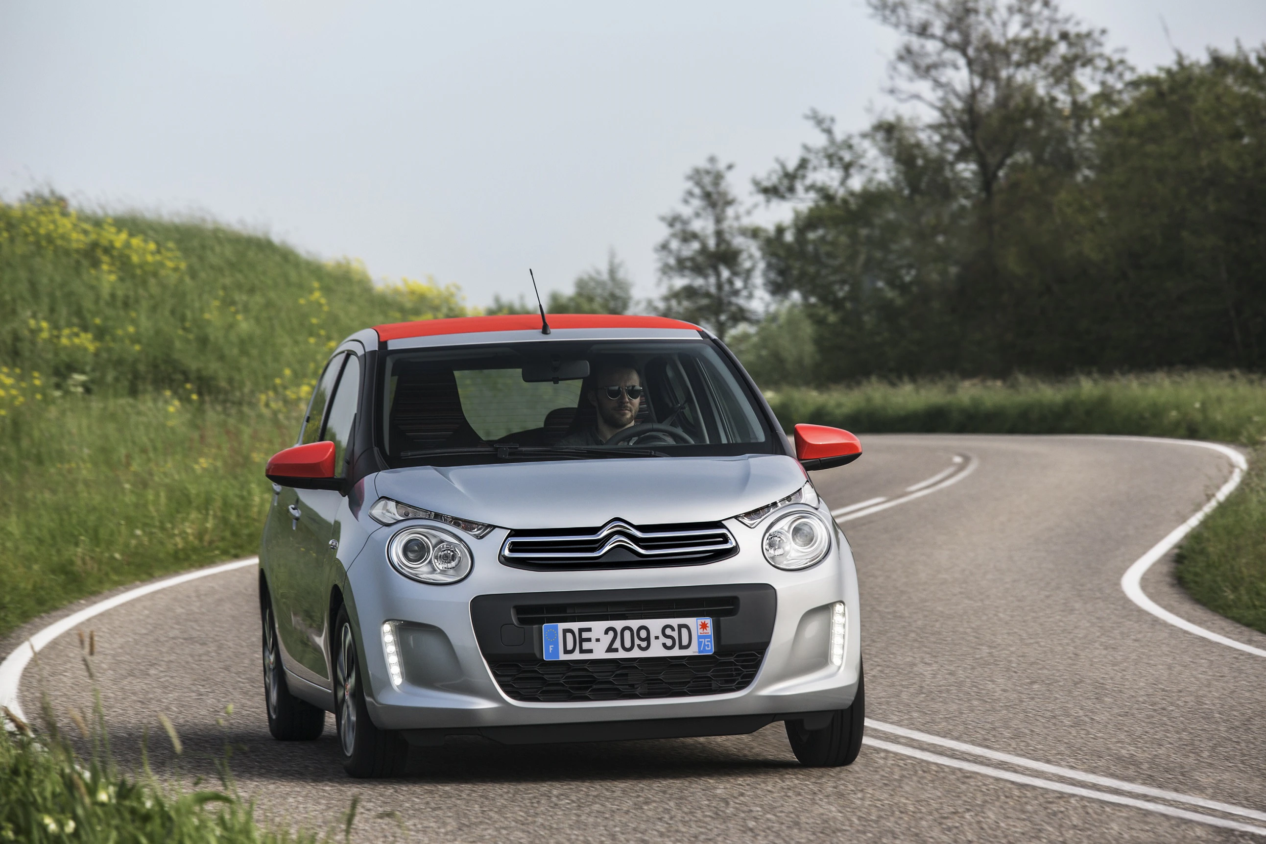 Silberner Citroën mit rotem Dach fährt auf einer kurvigen Landstraße durch grüne Landschaft. Fahrer trägt Sonnenbrille.