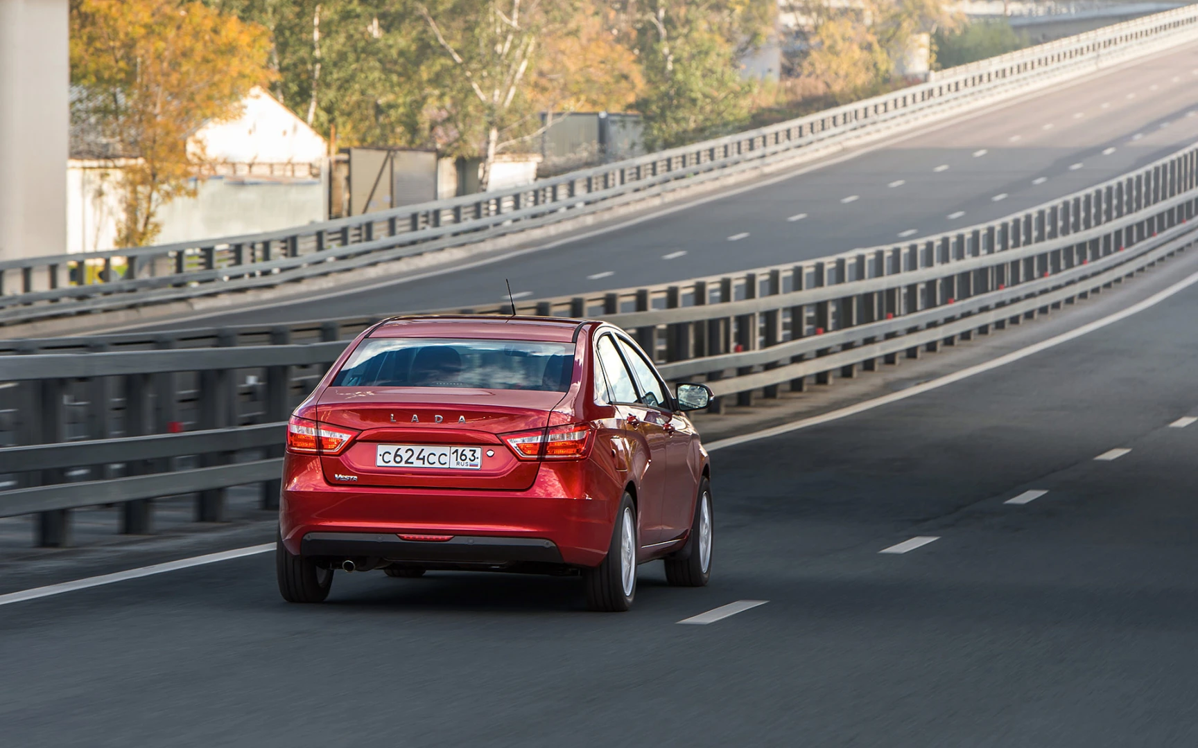 Rotes Auto fährt auf leerer, zweispuriger Autobahn mit Leitplanken, umgeben von Herbstbäumen. Lizenzplatte sichtbar.
