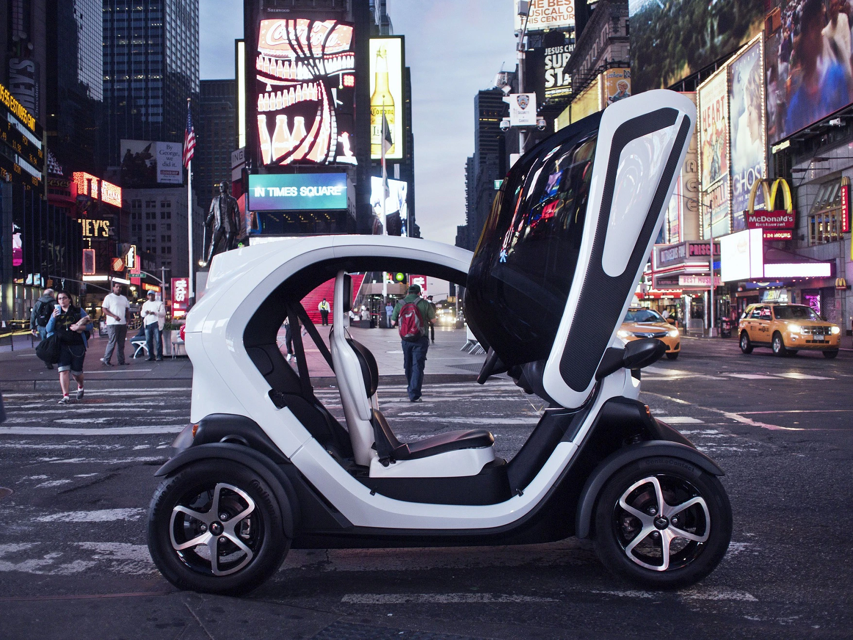 Kleines, futuristisches Elektroauto mit geöffneten Flügeltüren steht bei Nacht auf einer belebten Straße voller leuchtender Reklametafeln.
