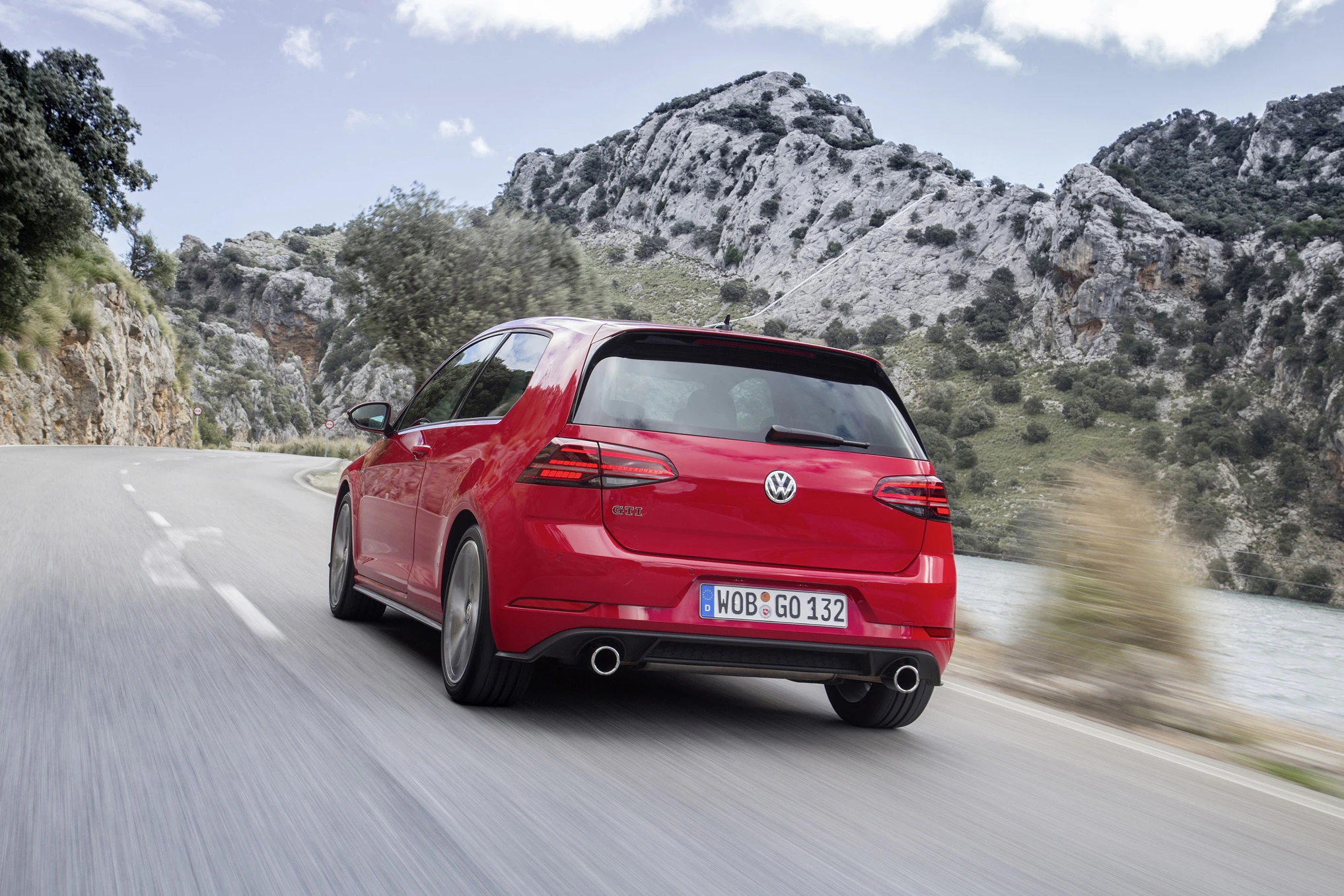 Roter VW Golf GTI fährt kurvige Bergstraße entlang, umgeben von Felsen und Bäumen, unter blauem Himmel.