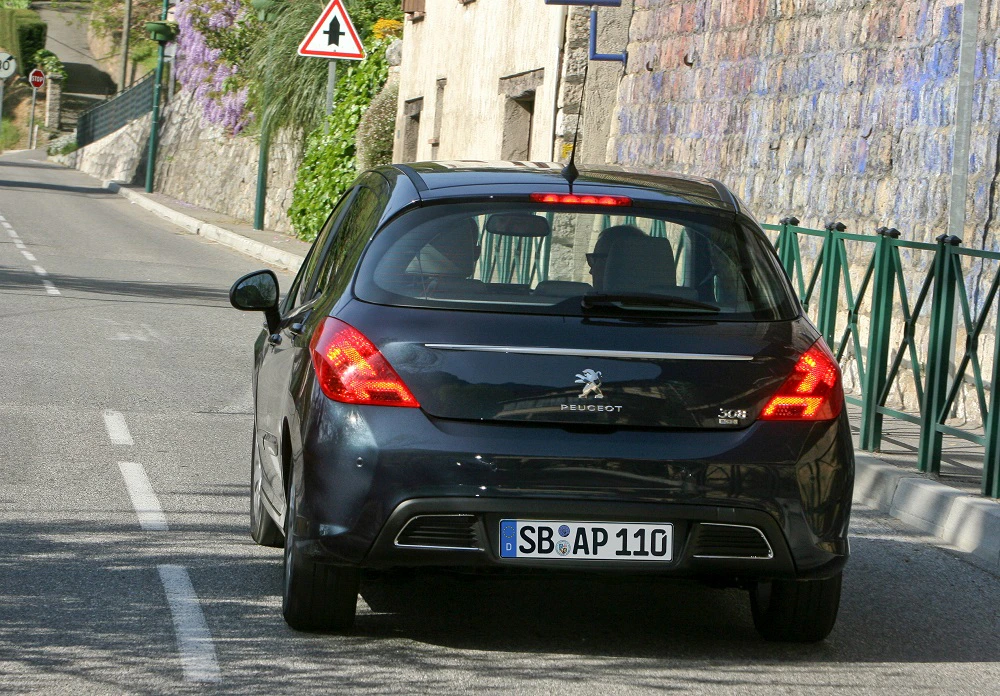 Schwarzer Peugeot 308 fährt auf schmaler, gepflasterter Straße mit Verkehrsschild und umgebenden Steinzäunen.
