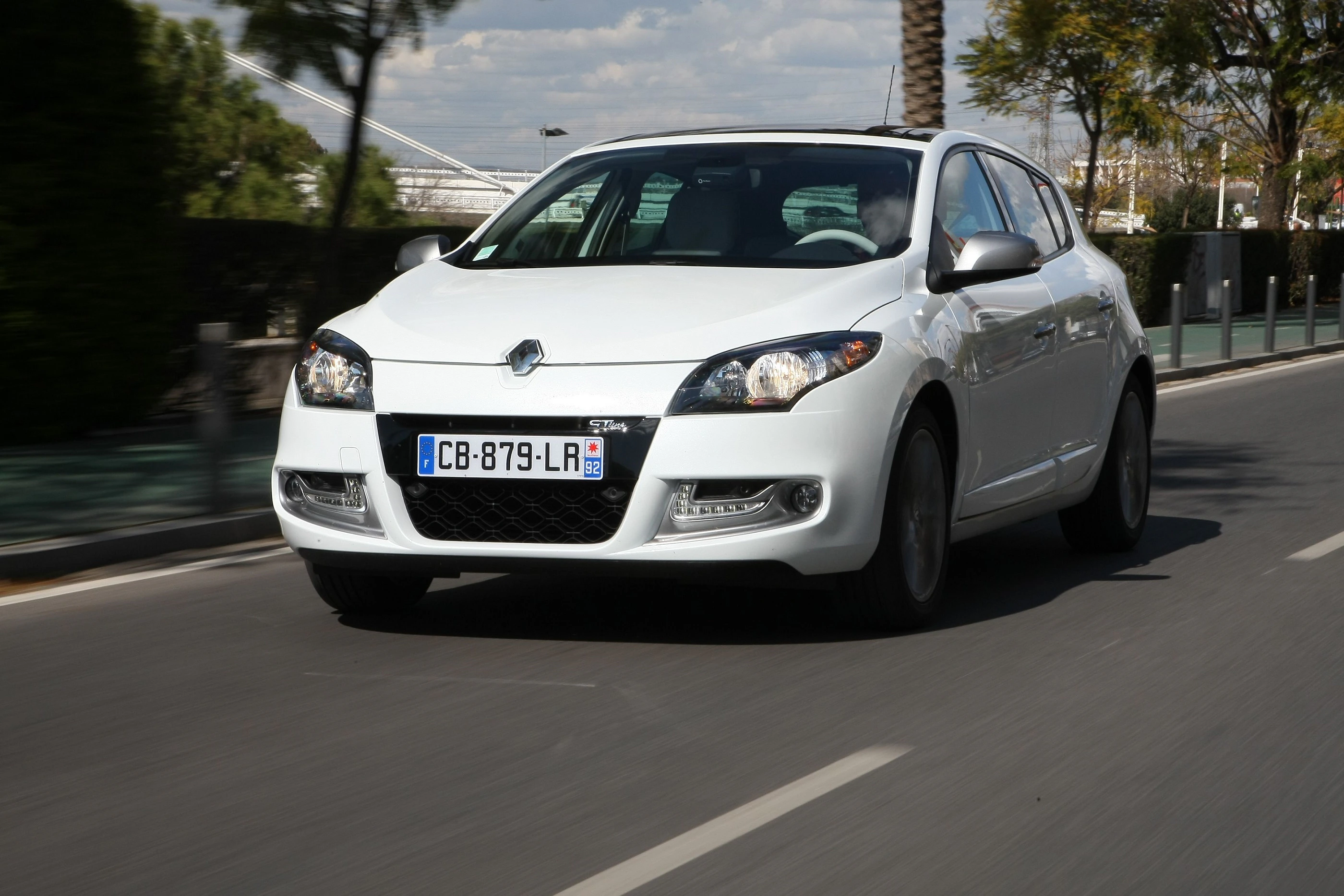 Weißer Renault Mégane fährt auf einer städtischen Straße, umgeben von Bäumen und moderner Architektur bei Tageslicht.