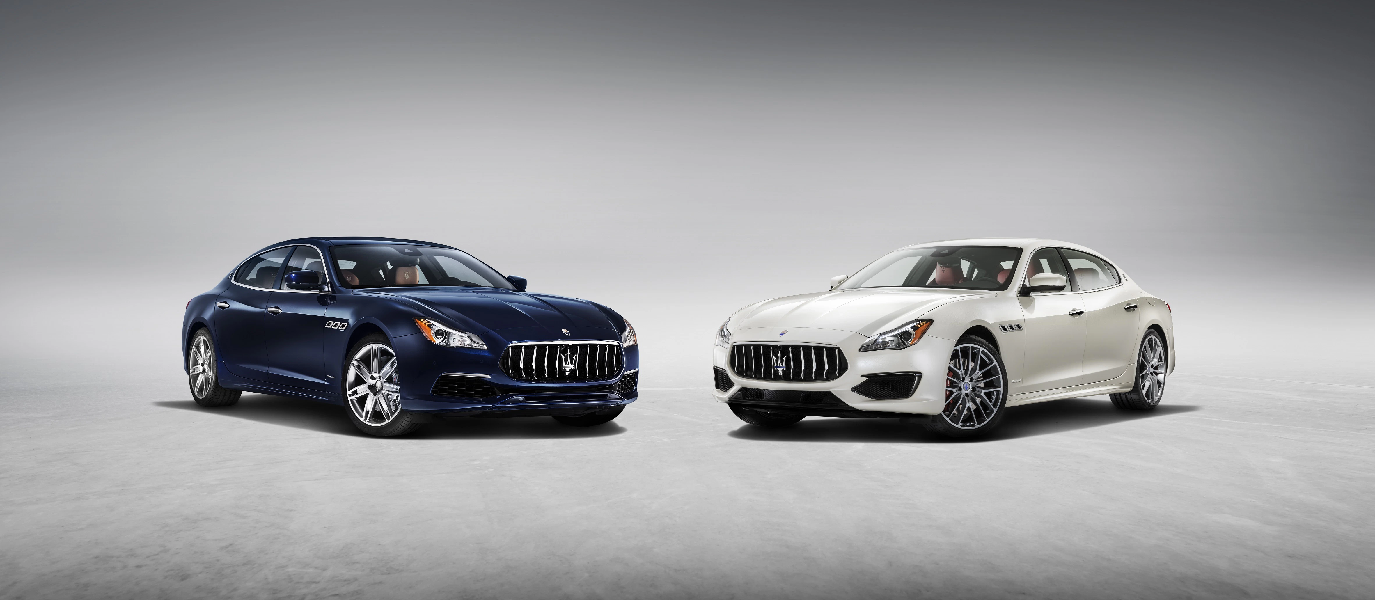 Zwei luxuriöse Maserati Limousinen, eine in Dunkelblau, die andere in Weiß, stehen nebeneinander auf hellem Boden vor grauem Hintergrund.