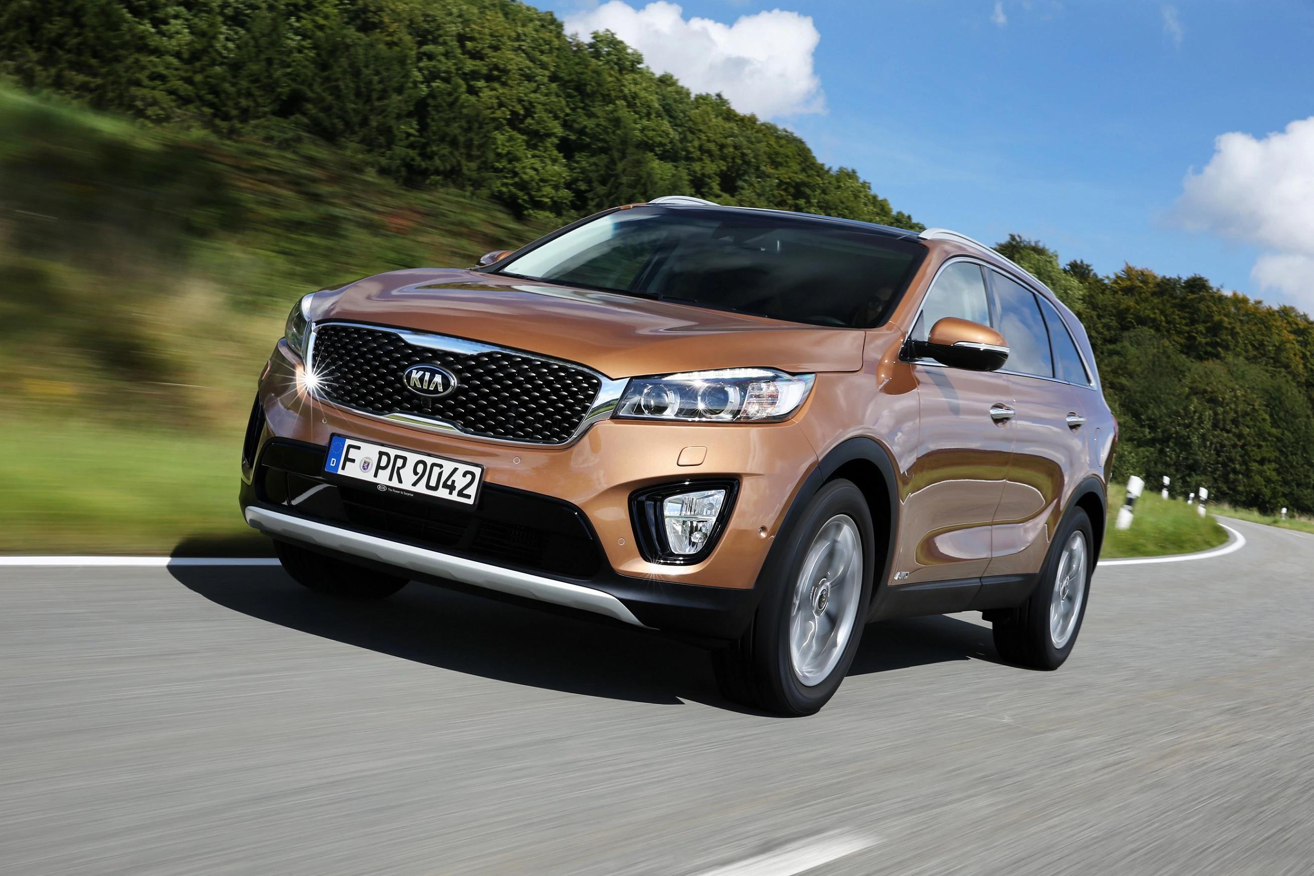 Brauner Kia SUV fährt auf einer kurvigen Landstraße bei klarem Himmel, im Hintergrund grüne Bäume. Fahrzeug mit deutschem Kennzeichen.