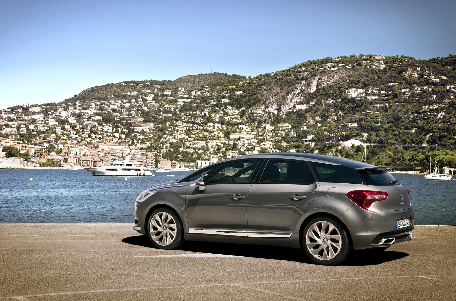 Silberner Citroën DS5 vor maritimer Kulisse mit blauem Wasser und bewaldeten Hügeln im Hintergrund. Yacht und Boote sind sichtbar.