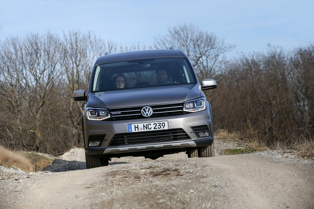 Silberner VW Caddy fährt auf steinigem Weg durch eine ländliche Landschaft, Bäume im Hintergrund. Zwei Personen sitzen im Auto.