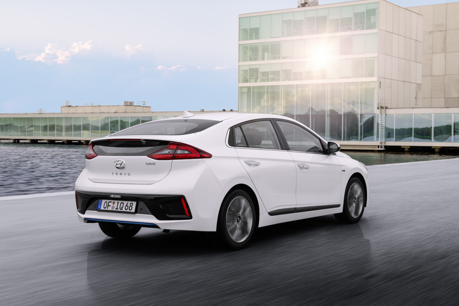 Weißer Hyundai Ioniq Hybrid fährt auf nasser Straße vor modernem Gebäude bei Sonnenschein.