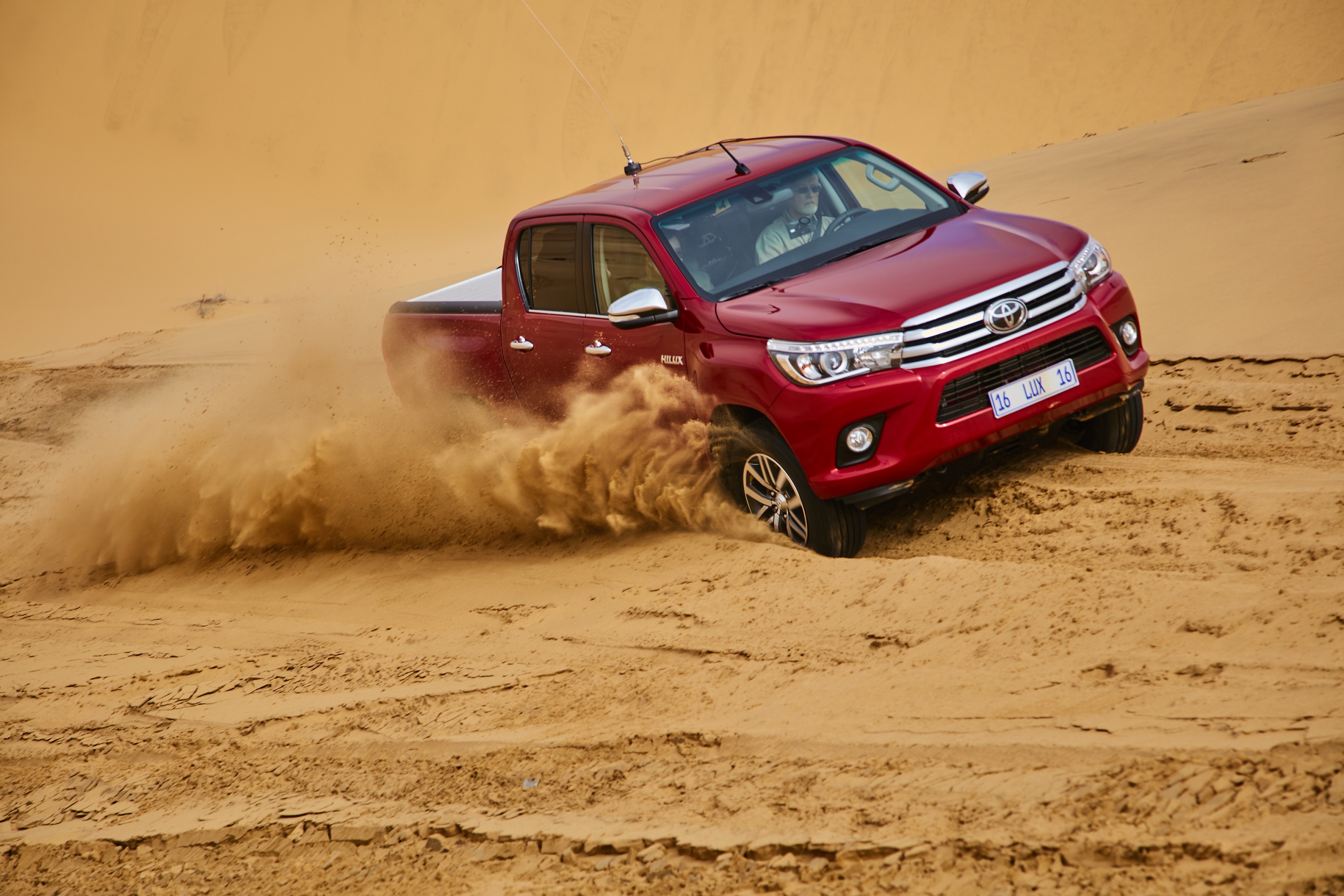Roter Pick-up fährt dynamisch durch Sanddünen, wirbelt Staub und Sand auf. Fahrzeug zeigt Leistungsfähigkeit im Offroad-Terrain.