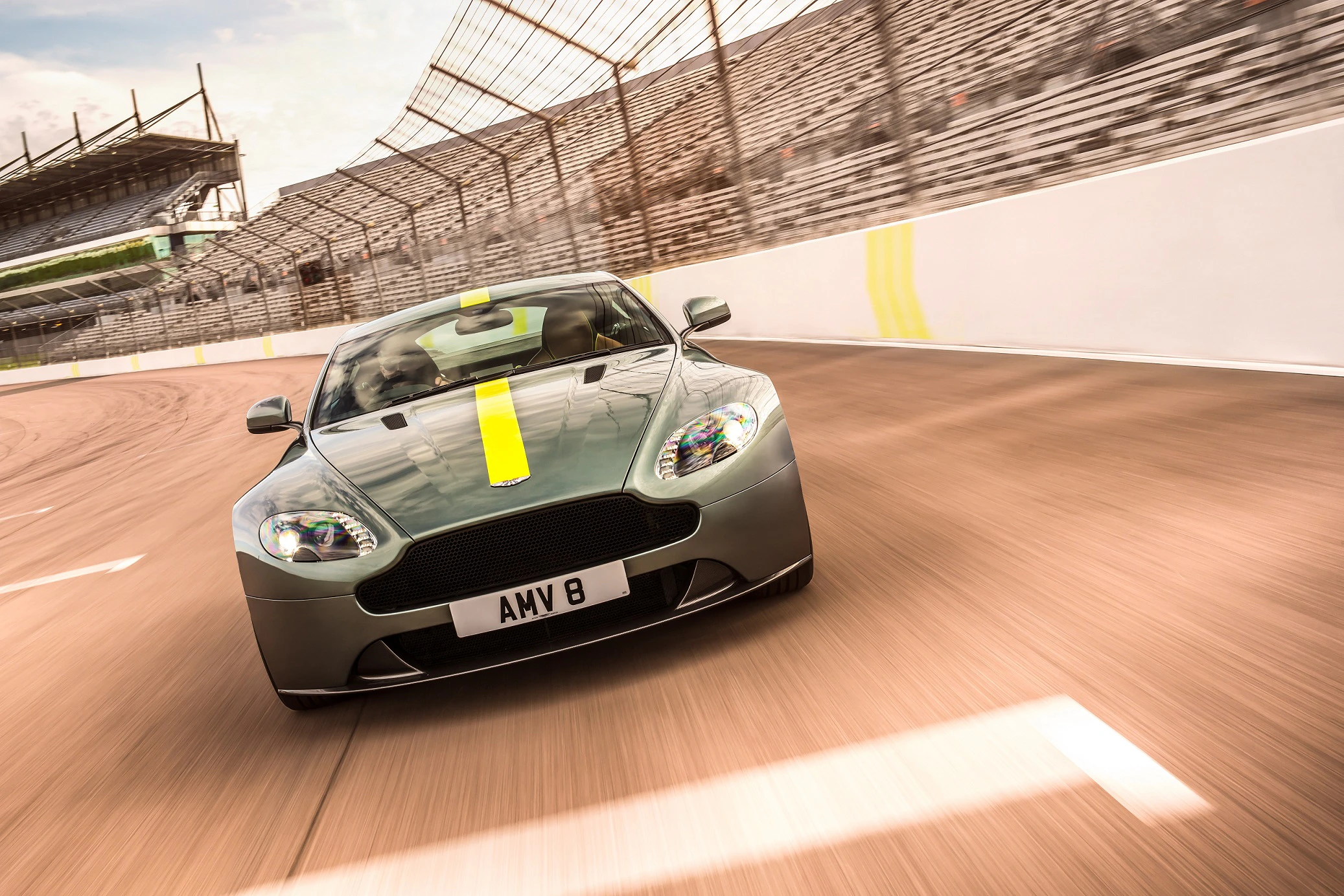 Aston Martin Vantage fährt mit hoher Geschwindigkeit auf Rennstrecke. Graues Auto mit gelbem Streifen, leere Tribünen im Hintergrund.