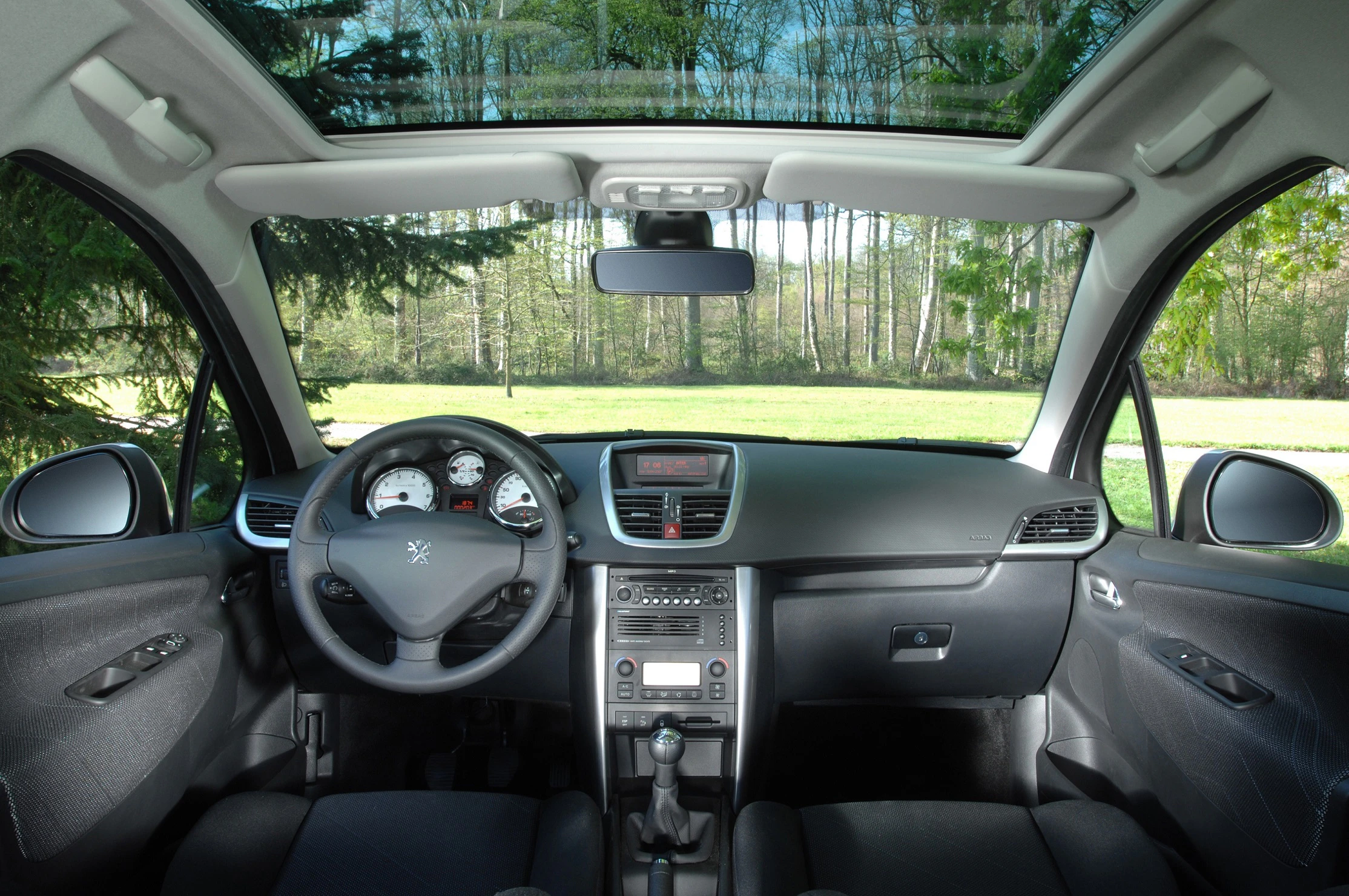 Innenraum eines Autos mit Glasdach, Lenkrad, Armaturenbrett und manuellem Getriebehebel. Außen ist ein Wald sichtbar.