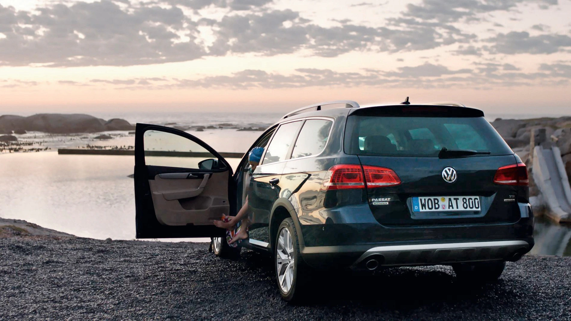 Ein Volkswagen Kombi parkt vor einem See bei Sonnenuntergang. Zwei Personen sitzen im Auto, eine Tür ist geöffnet.