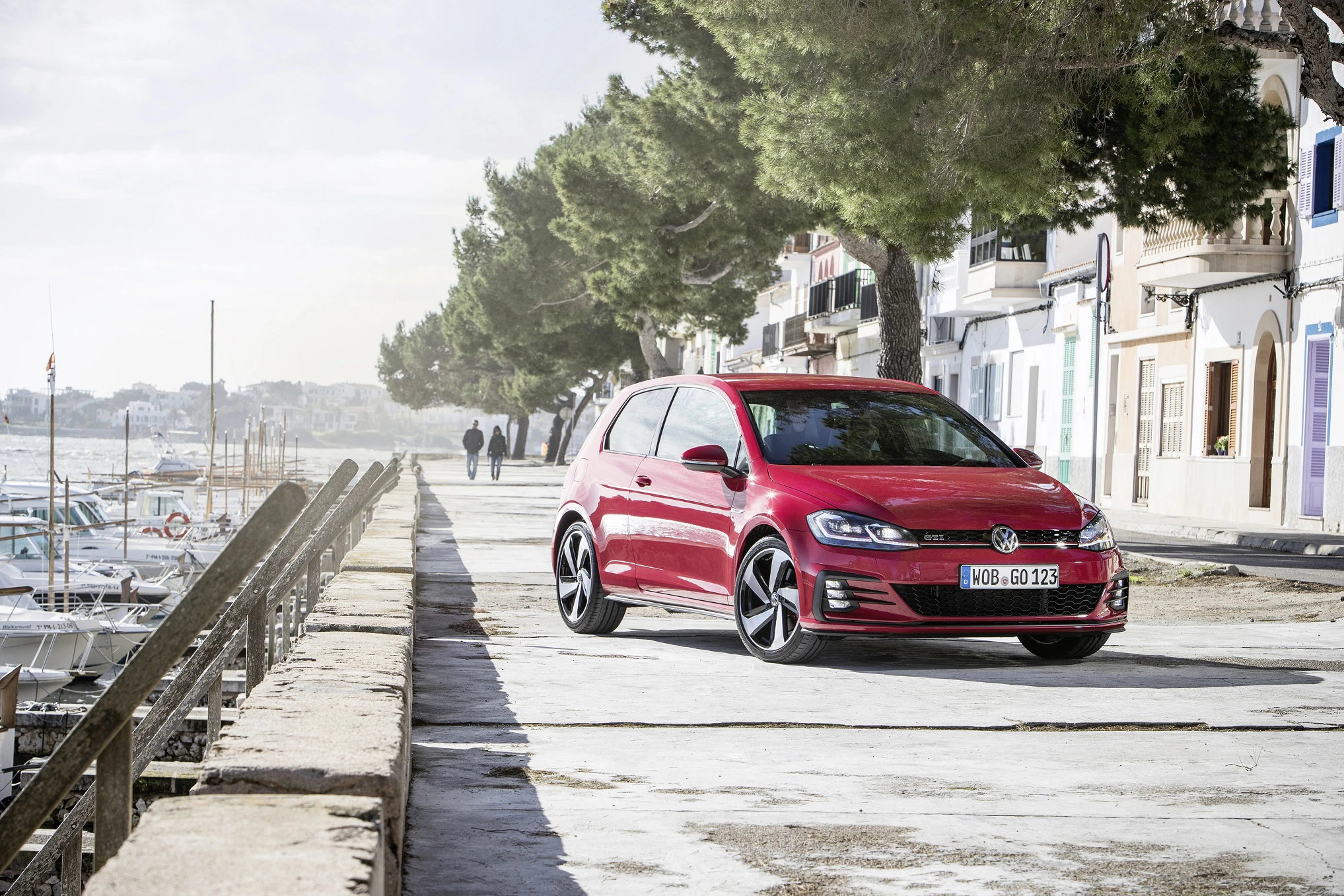 Roter VW Golf fährt auf einer Küstenstraße entlang, im Hintergrund Bäume und Gebäude, daneben das Meer mit Booten.