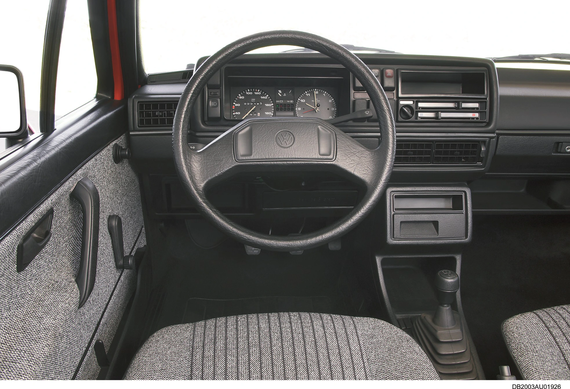 Innenraum eines klassischen Autos mit schwarzem Lenkrad, Armaturenbrett und grauen Sitzbezügen. Anzeigeinstrumente und Schaltknauf sichtbar.