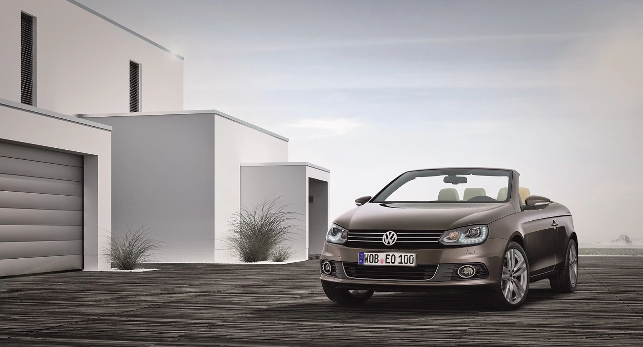 Braunes VW-Cabrio geparkt vor modernem, weißem Gebäude auf Holzterrasse, bewölkter Himmel im Hintergrund.