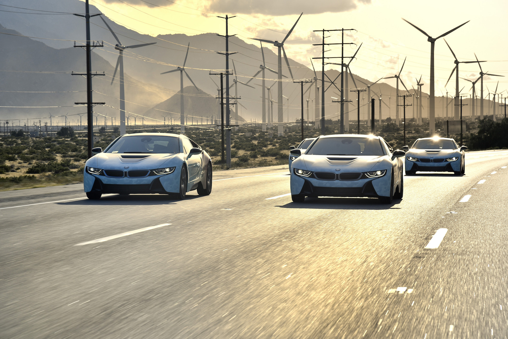 Drei silberne Sportwagen fahren auf einer Straße in der Wüste. Im Hintergrund sind viele Windkraftanlagen unter einem bewölkten Himmel zu sehen.