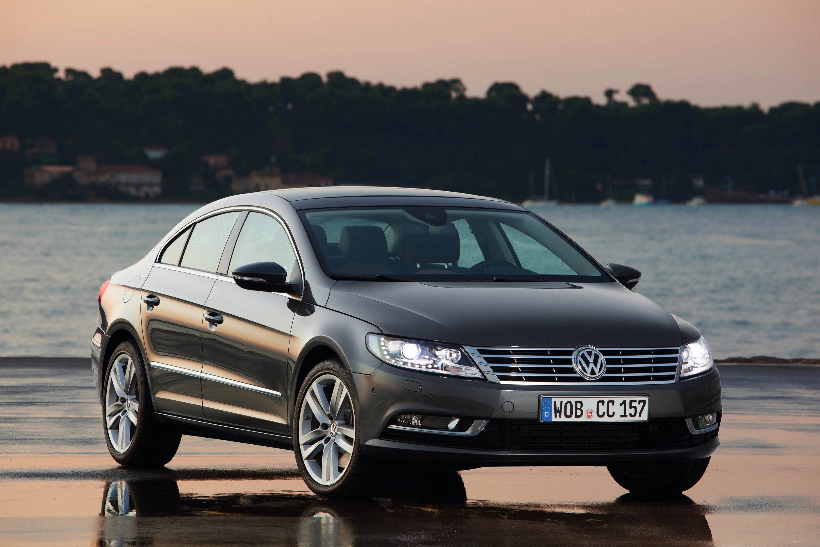 Silberner Volkswagen CC parkt bei Sonnenuntergang an einem Gewässer mit Bäumen im Hintergrund.