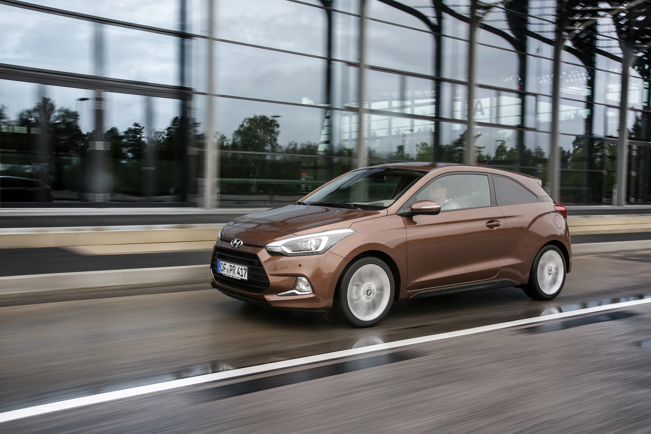 Brauner Hyundai i20 fährt auf nasser Straße vor moderner Glasfassade. Subtiler Bewegungseffekt auf Rädern. Bewölkter Himmel spiegelt sich im Glas.