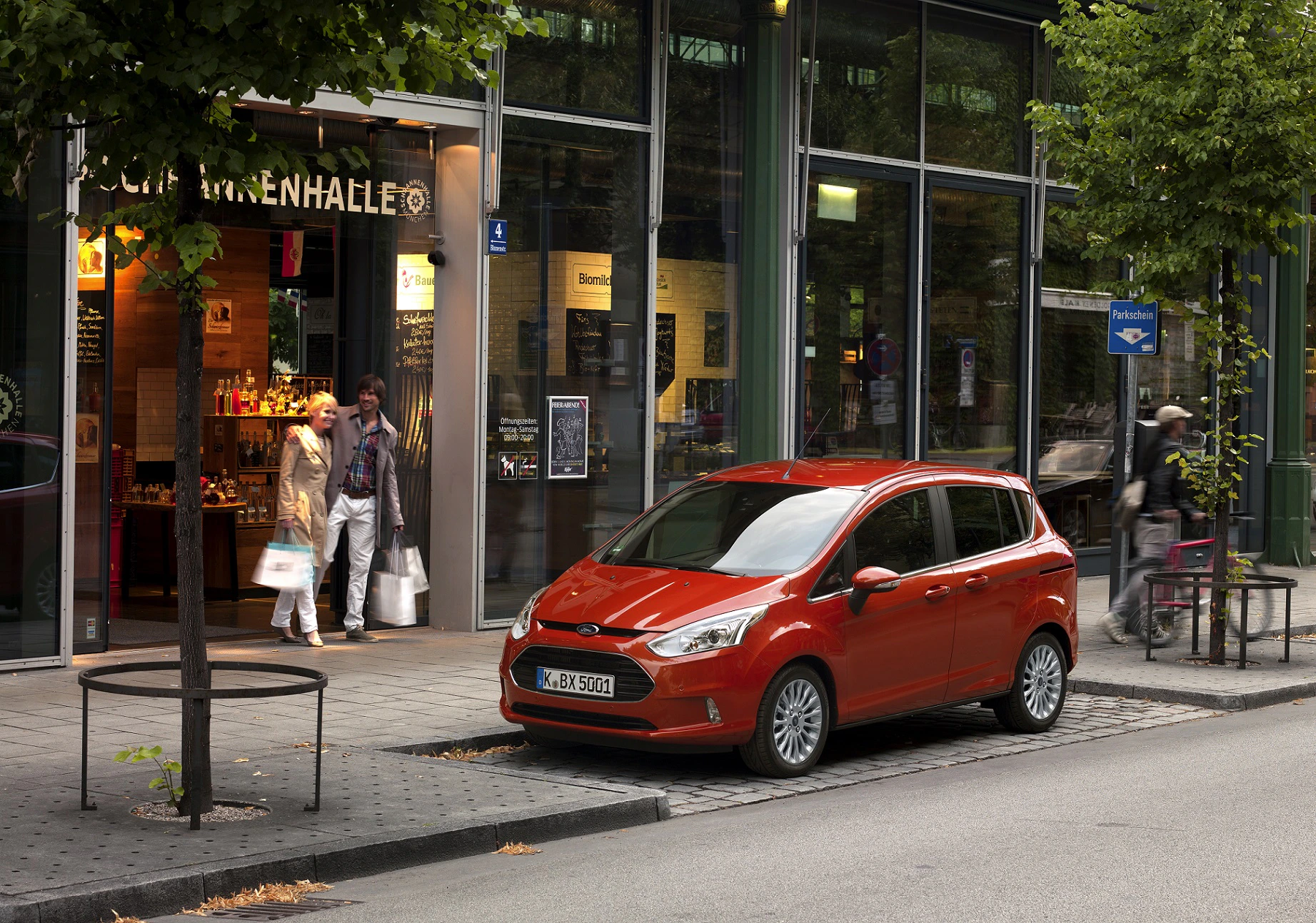Rotes Auto parkt vor einem Geschäft mit Personen am Eingang; grüne Bäume und ein blaues Verkehrsschild daneben.