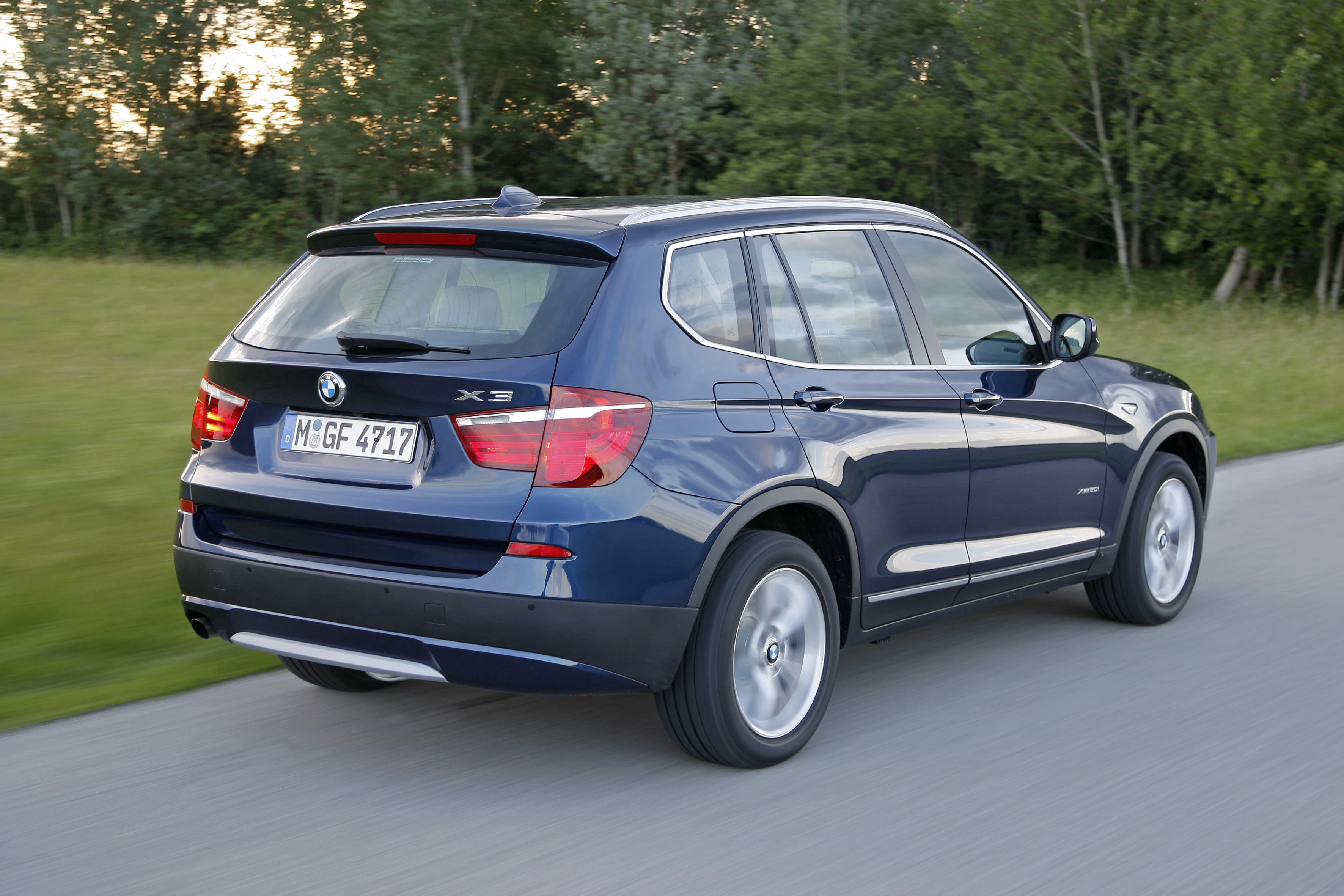 Ein blauer BMW X3 fährt auf einer Landstraße, umgeben von grünen Bäumen und Wiesen.
